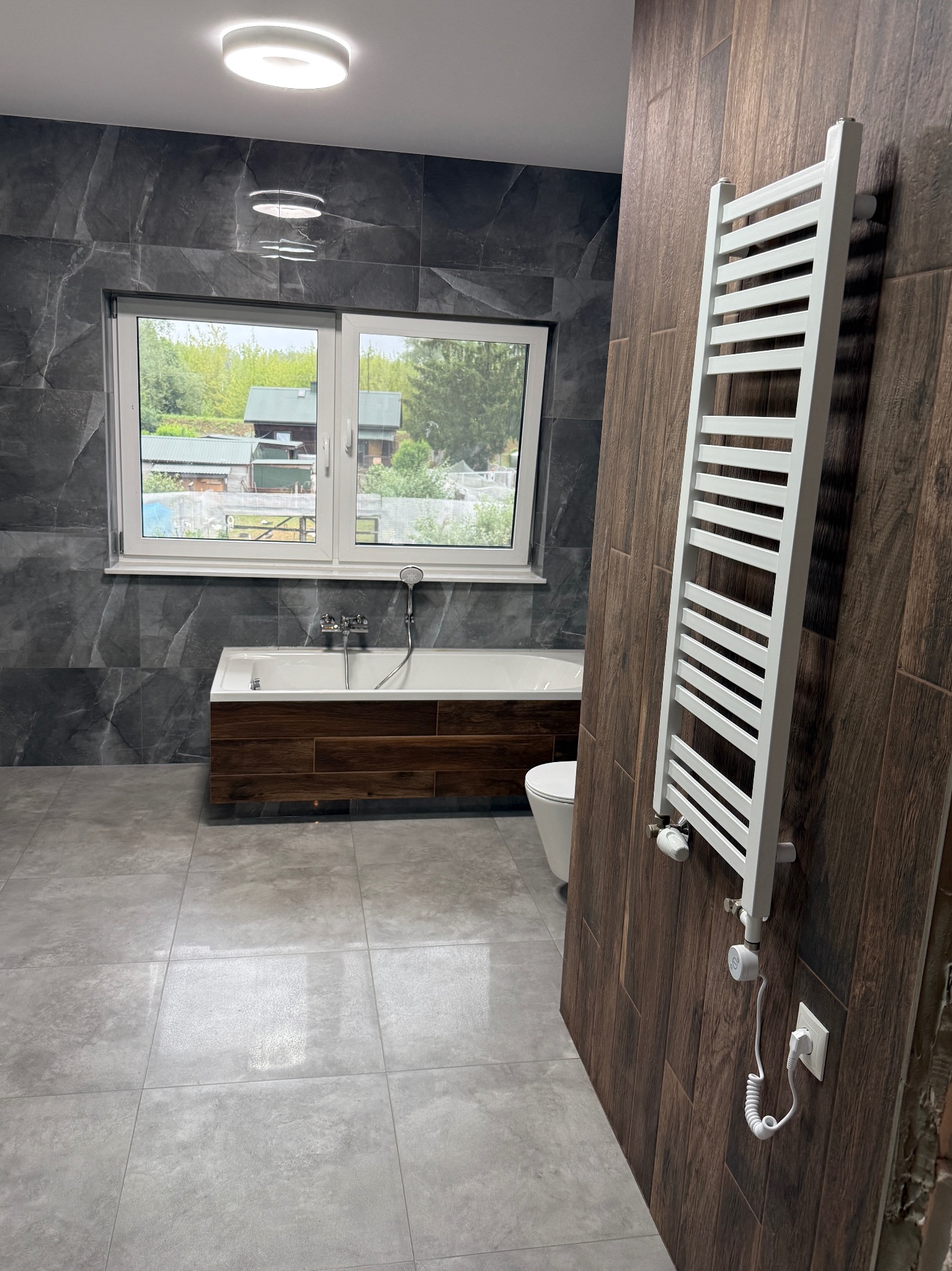 Nowoczesna łazienka z szarymi płytkami imitującymi marmur i drewnianymi akcentami. Widoczna wanna, grzejnik drabinkowy i okno z widokiem na zieleń.