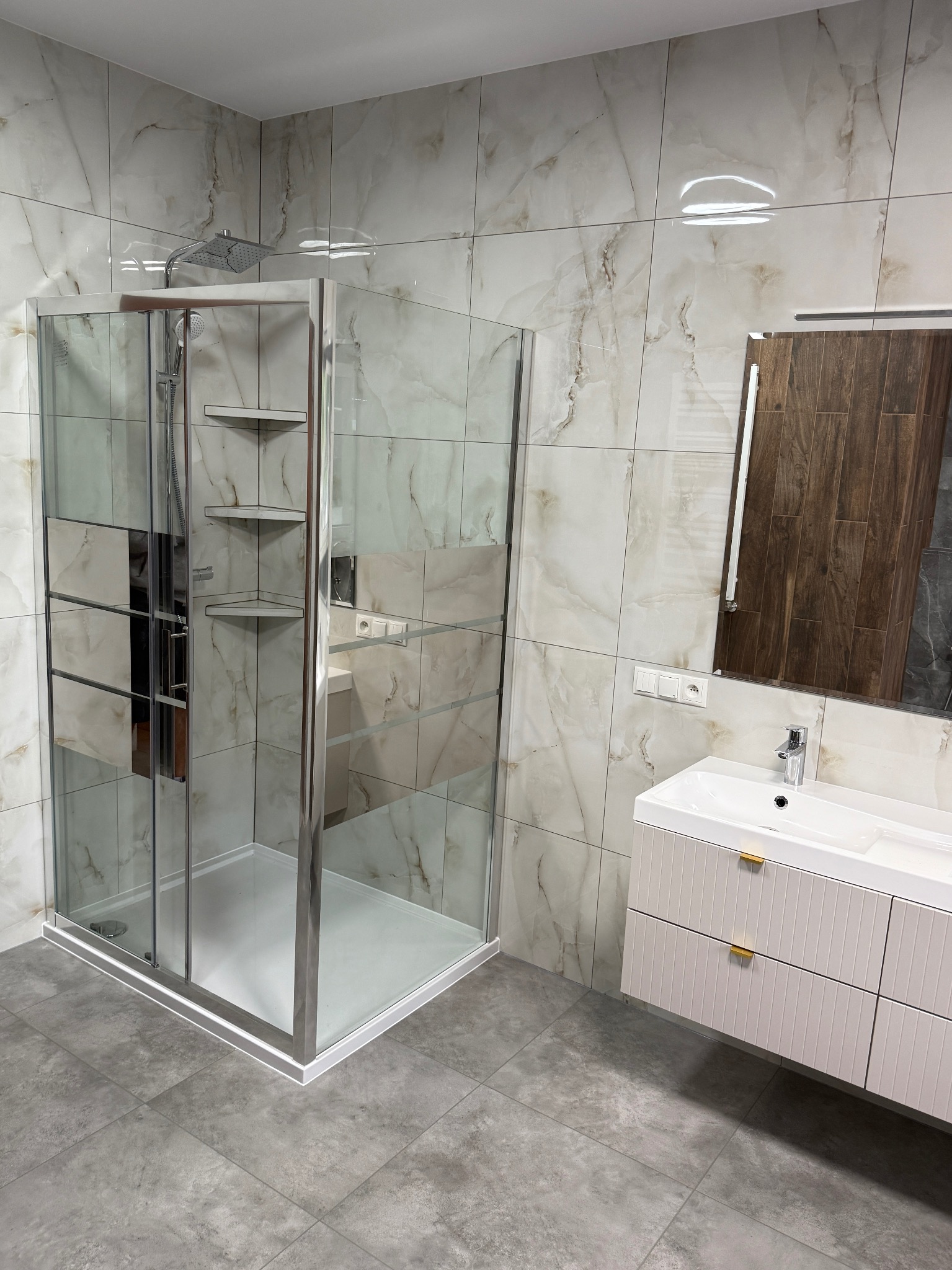 Nowoczesna łazienka z kabiną prysznicową typu walk-in, umywalką z szafką w kolorze złamanej bieli i lustrem w drewnianej ramie. Marmurowe płytki na ścianach i szare na podłodze.