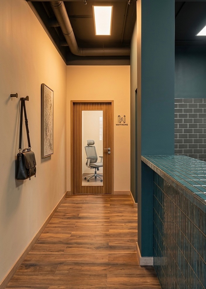 Nowoczesny korytarz biurowy z drewnianą podłogą, beżowymi ścianami, turkusową ladą i widokiem na przeszklone biuro z krzesłem obrotowym. Minimalistyczny design.