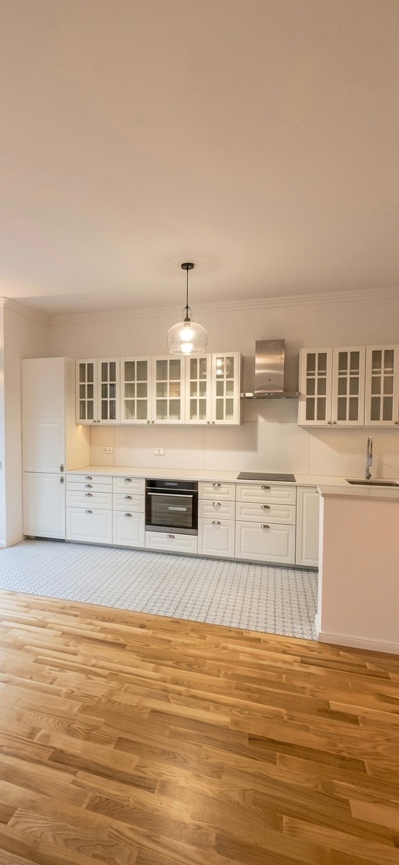 Jasna kuchnia z białymi szafkami, wzorzystą podłogą i drewnianym parkietem. Widok na całe pomieszczenie, z wiszącą lampą i srebrnym okapem.