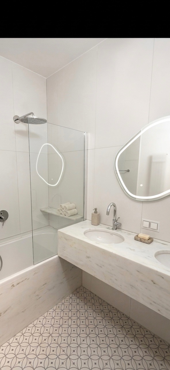 Nowoczesna łazienka w bieli i szarości. Umywalka z marmurowym blatem, lustro z podświetleniem LED i wzorzysta podłoga. Kabina prysznicowa ze szklaną ścianką.