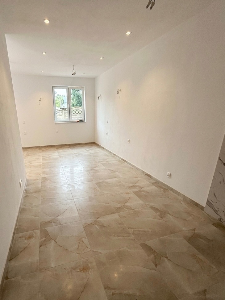 Jasne wnętrze po remoncie: beżowe płytki podłogowe z marmurkowym wzorem, białe ściany z widocznymi gniazdkami i oknem. Minimalistyczny design, gotowe do umeblowania.
