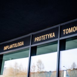 DentistiQ - Zewnętrzny widok nowoczesnego budynku z ciemną fasadą i dużymi oknami, nad którymi widnieją złote napisy: 'SKANER', 'IMPLANTOLOGIA', 'PROTETYKA', 'TOMOGRAFIA'.