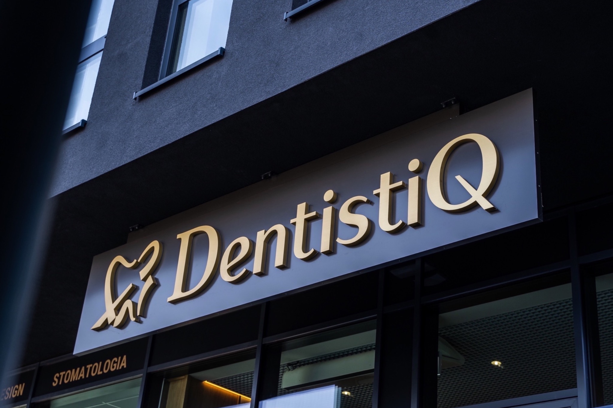 Złoty szyld z nazwą 'DentistiQ' i symbolem zęba w kształcie serca na ciemnoszarym budynku kliniki.