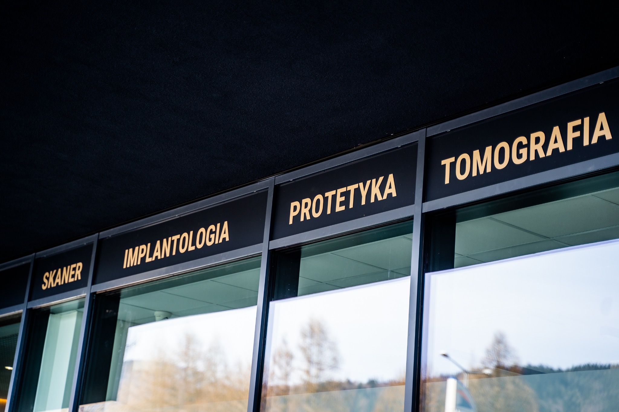 Zewnętrzny widok okien placówki medycznej z napisami w kolorze złotym: Skaner, Implantologia, Protetyka, Tomografia.
