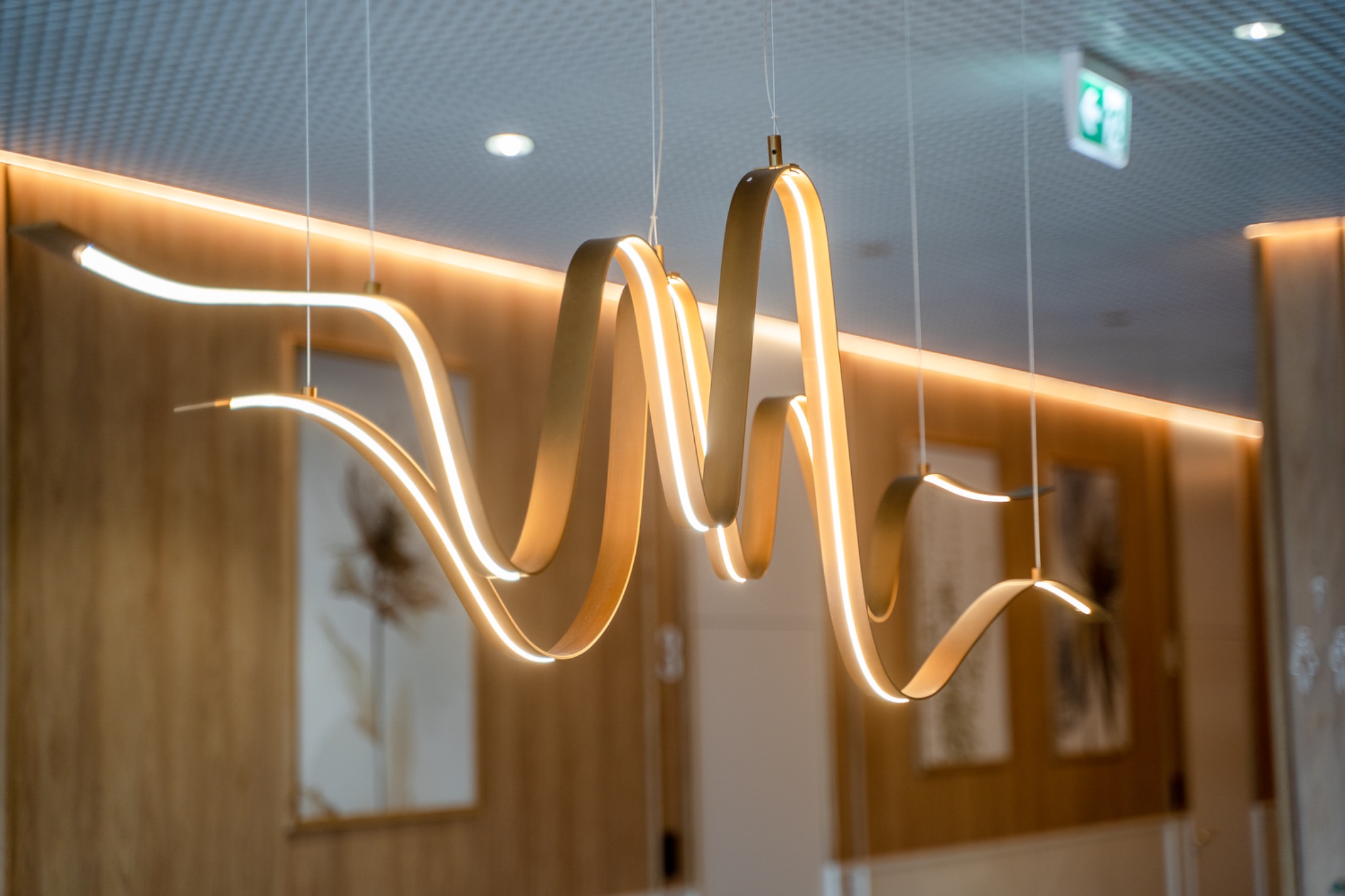 Nowoczesna lampa wisząca z falistymi, złotymi elementami LED w oświetlonym wnętrzu z drewnianymi panelami i obrazami w tle.