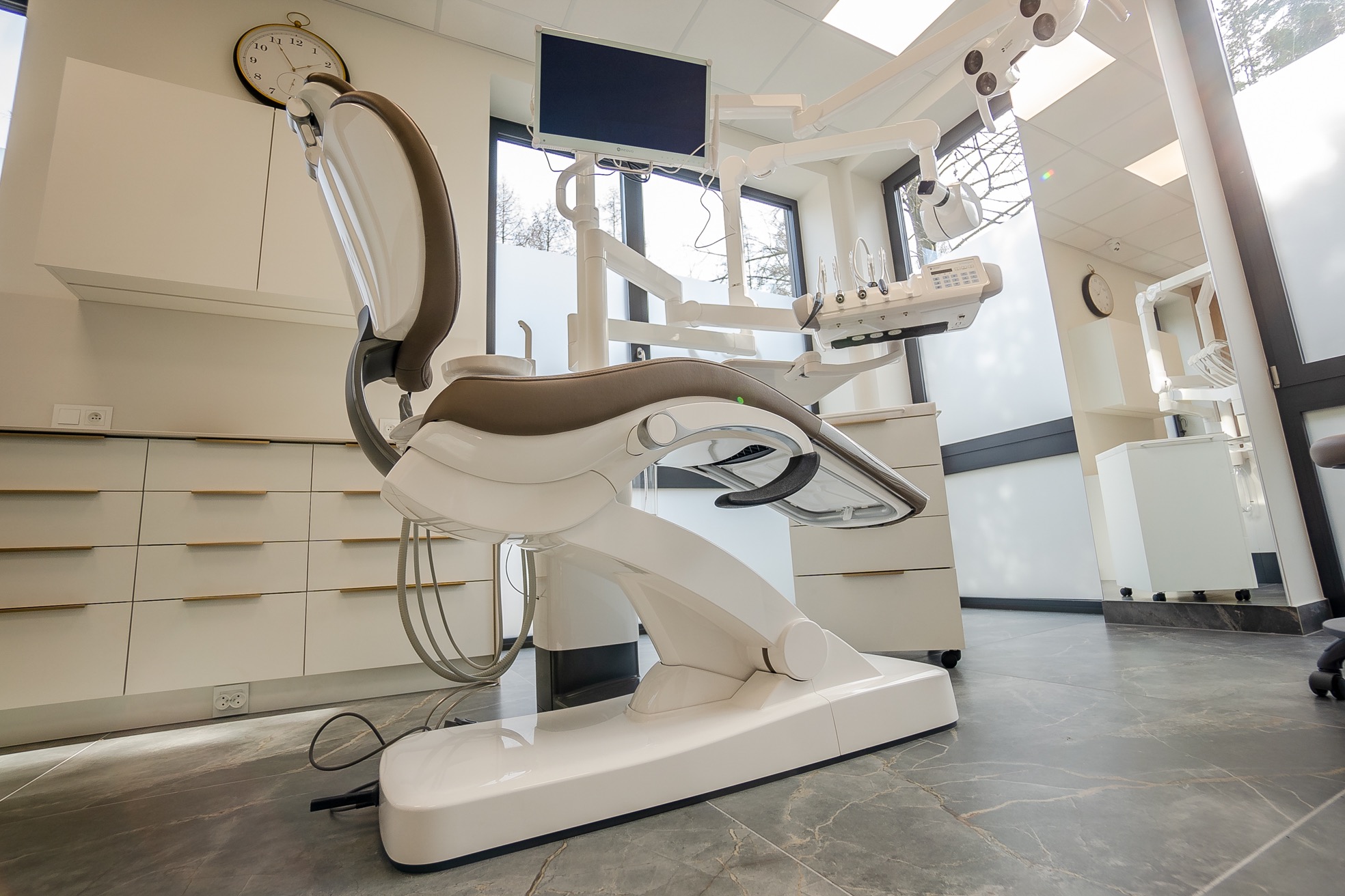 Nowoczesny, biało-szary fotel dentystyczny z ekranem i ramieniem z narzędziami w jasnym gabinecie, widok z boku, na ścianie zegar.