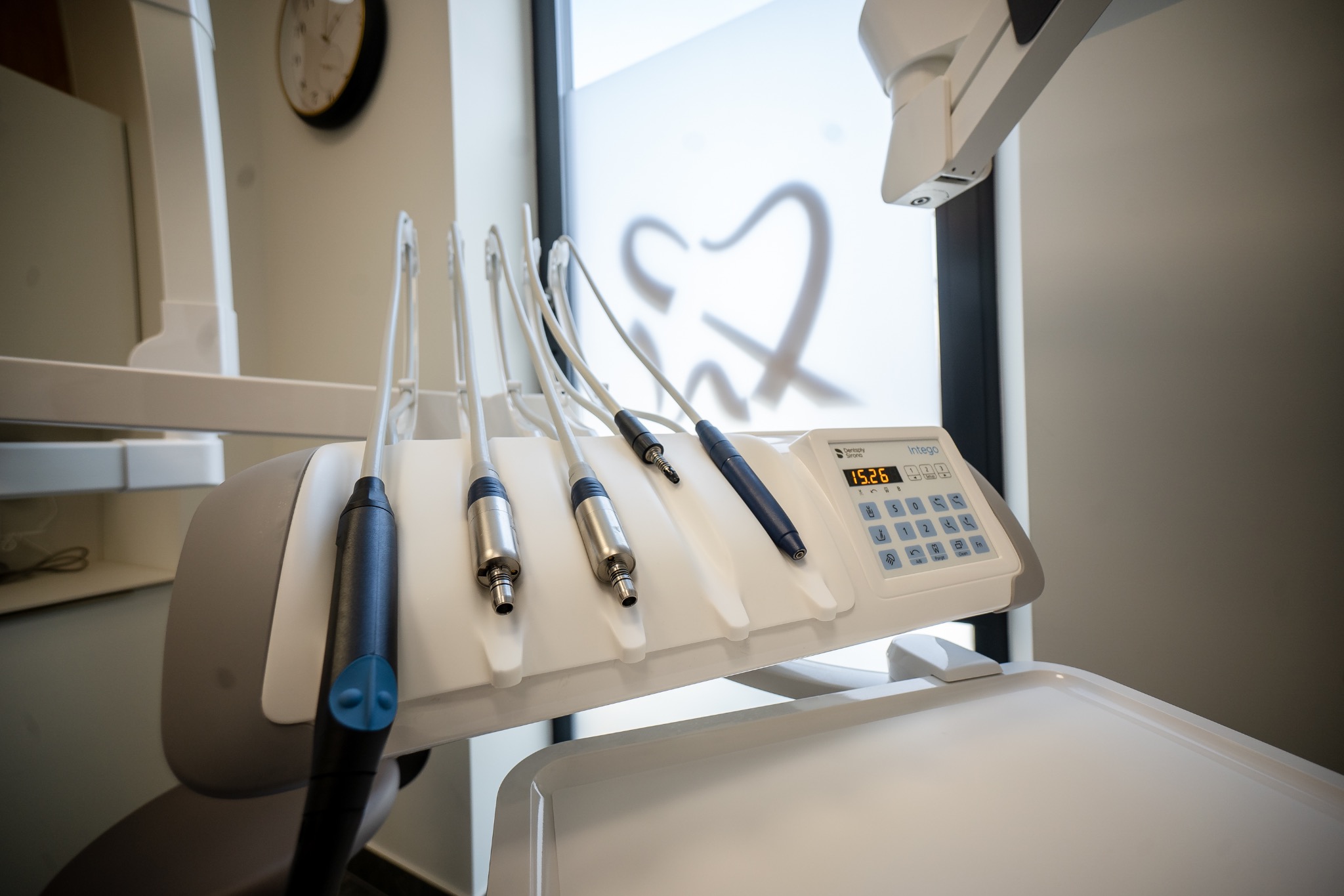 Nowoczesny unit zabiegowy z czterema końcówkami i panelem sterowania Intego Dentsply Sirona, w tle okno z motywem dentystycznym i zegar.