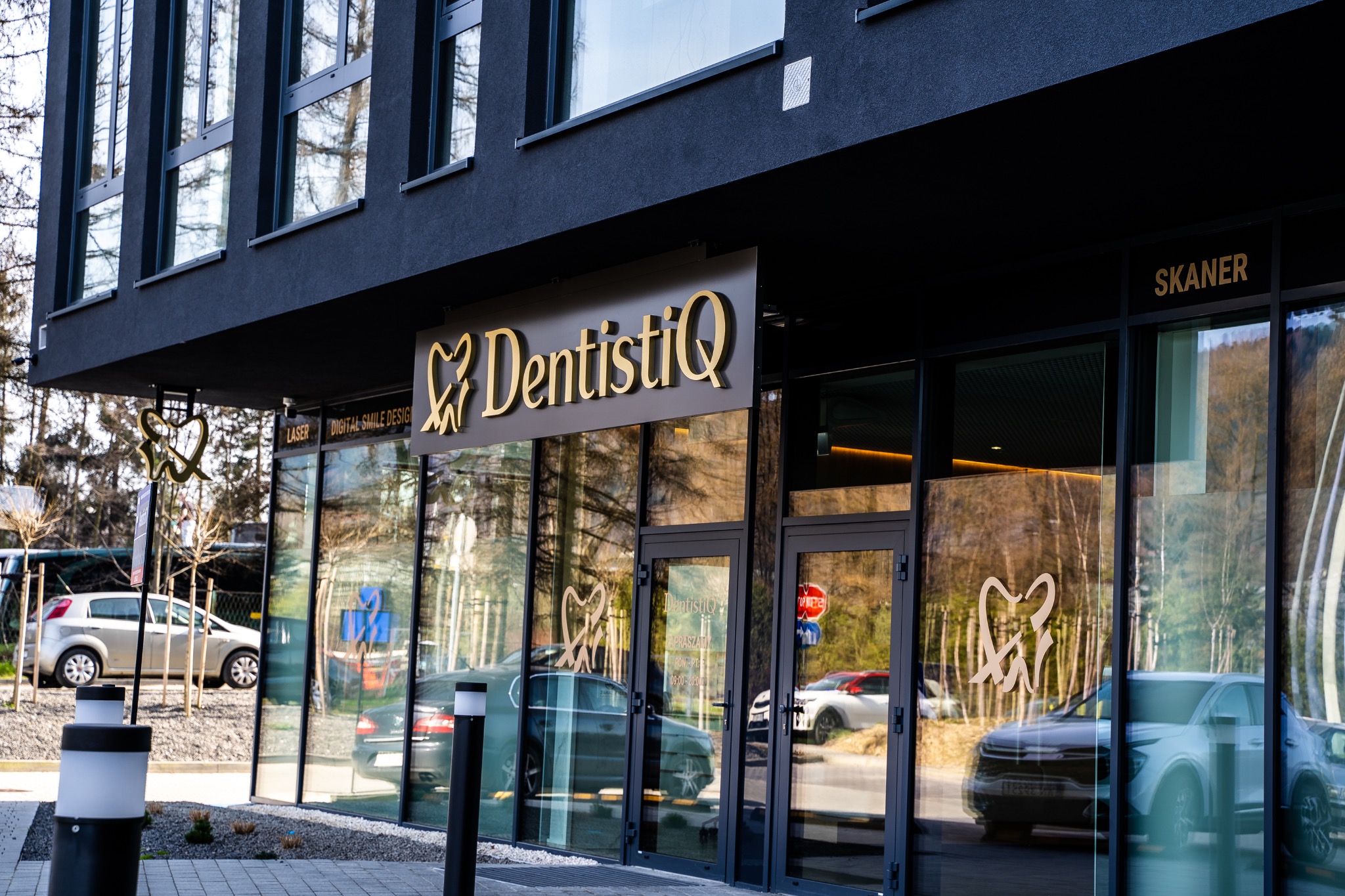 Elewacja nowoczesnej kliniki dentystycznej DentistIQ z dużym złotym logo i przeszklonymi ścianami, na których widnieją napisy 'Laser' i 'Digital Smile Design', z zaparkowanymi samochodami przed...