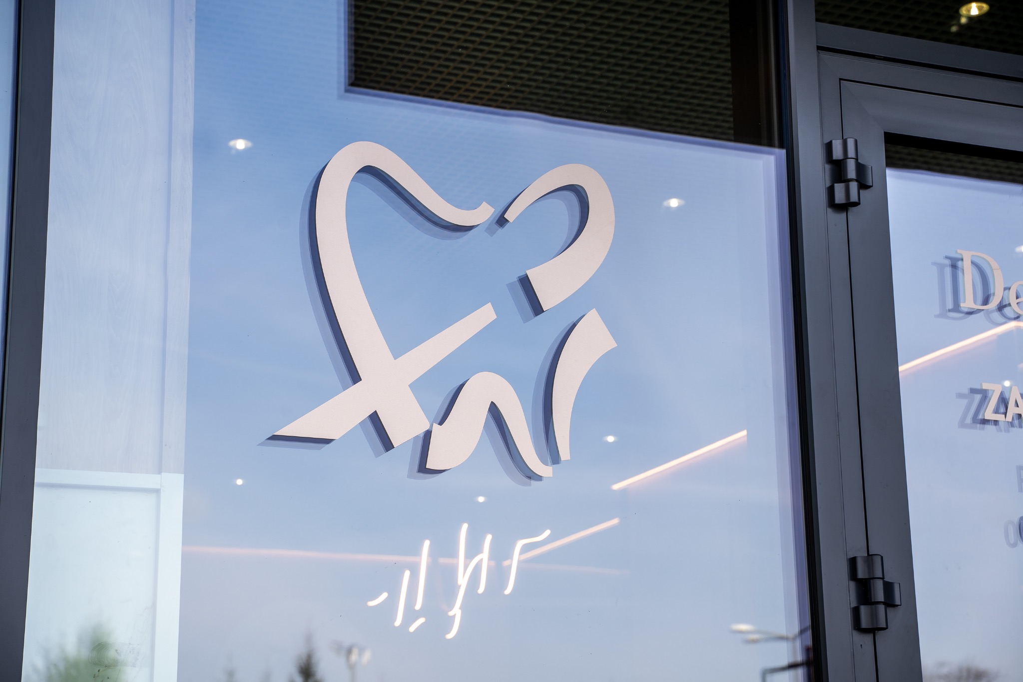 Stylizowane logo zęba na szklanej fasadzie budynku z ciemną stolarką okienną i odblaskami neonów.