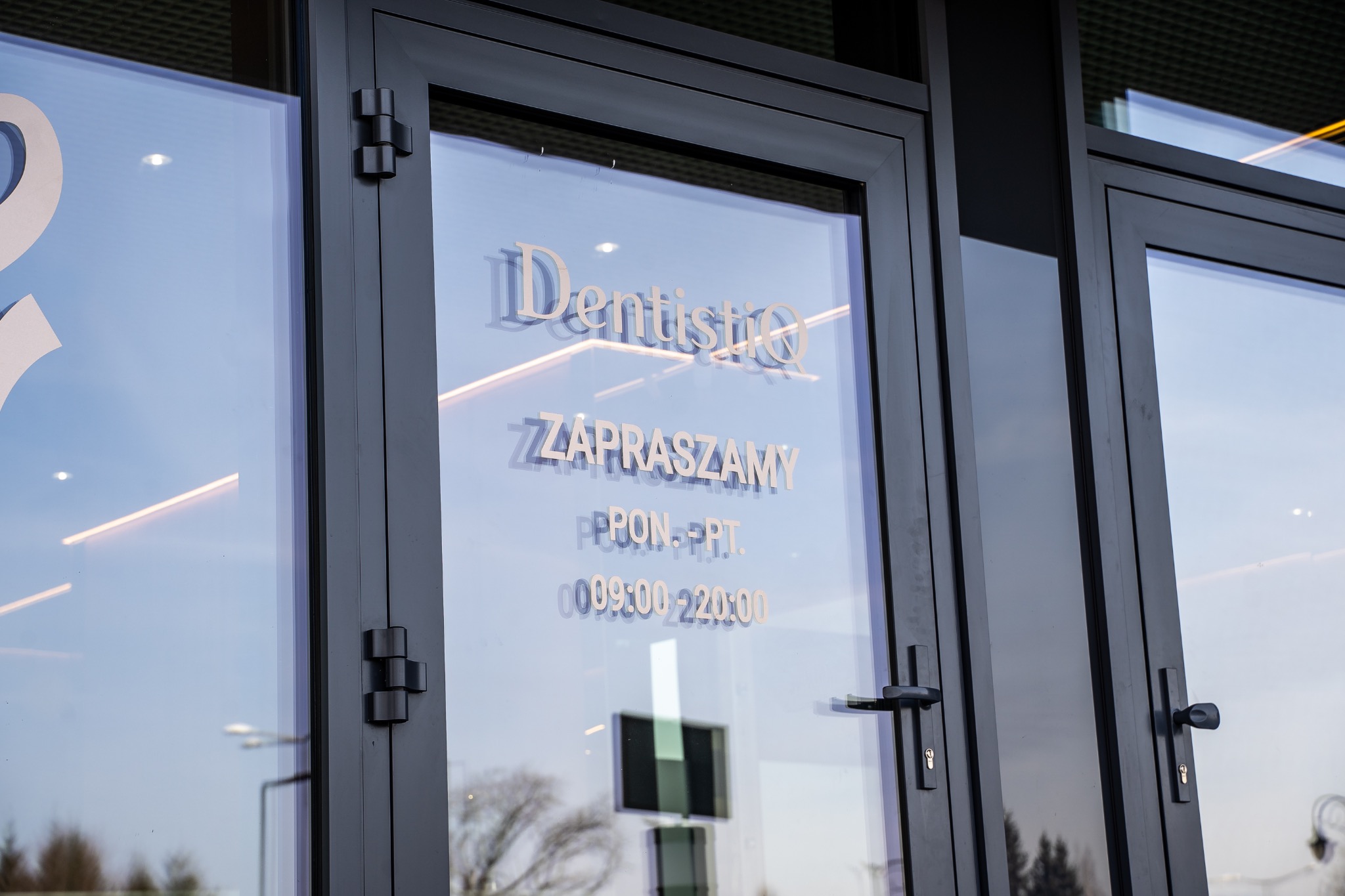 Wejście do kliniki dentystycznej 'DentistiQ' z godzinami otwarcia wyklejonymi na szklanych drzwiach, widoczne metalowe zawiasy i klamka.