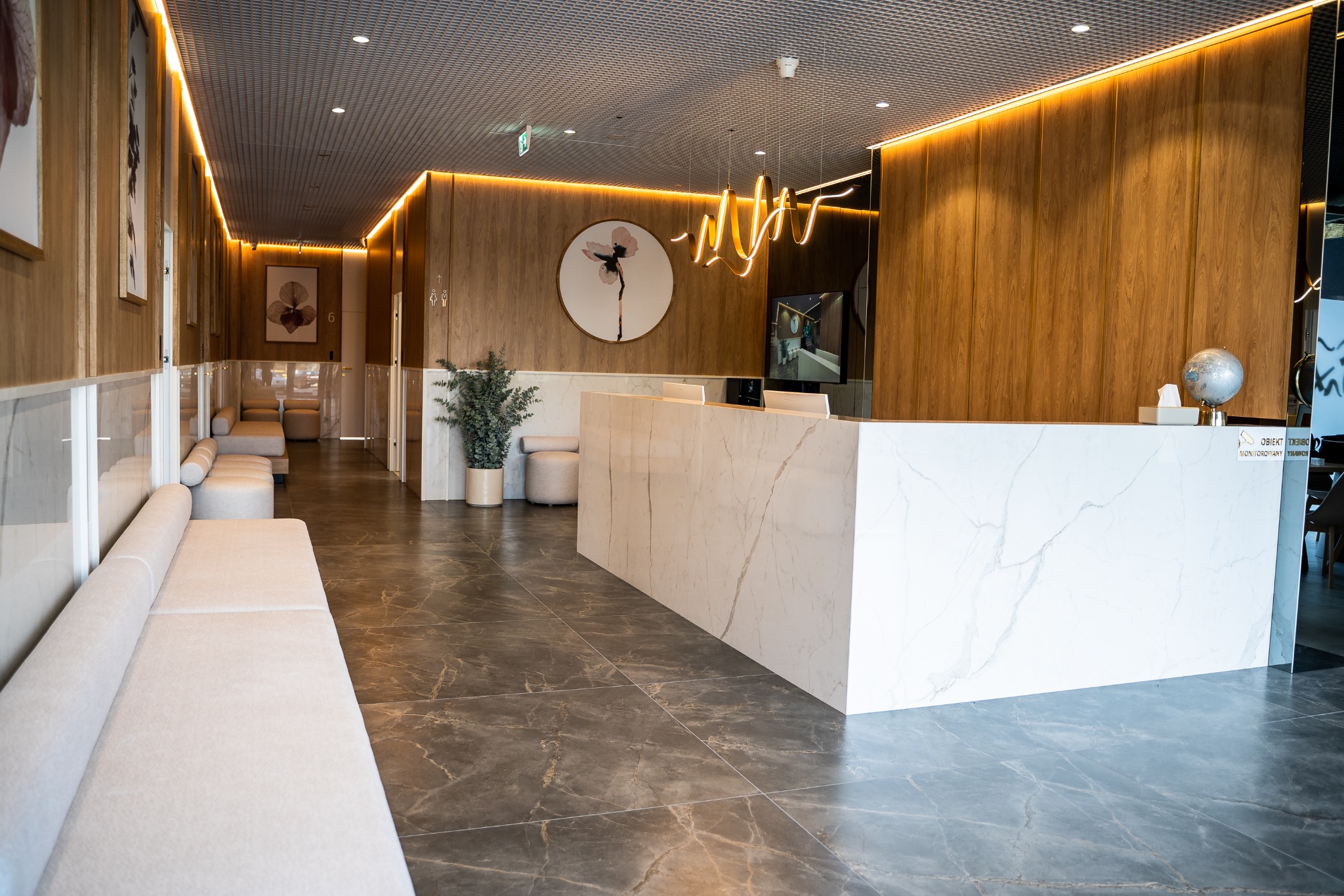Jasna recepcja z marmurowym blatem, drewnianymi panelami ściennymi z oświetleniem LED, nowoczesną lampą i rośliną doniczkową w eleganckim wnętrzu.