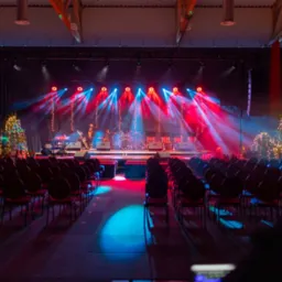 Scena przygotowana do występu z instrumentami, perkusją, rzędami krzeseł dla publiczności i dekoracjami świątecznymi, oświetlona czerwonymi i niebieskimi reflektorami.