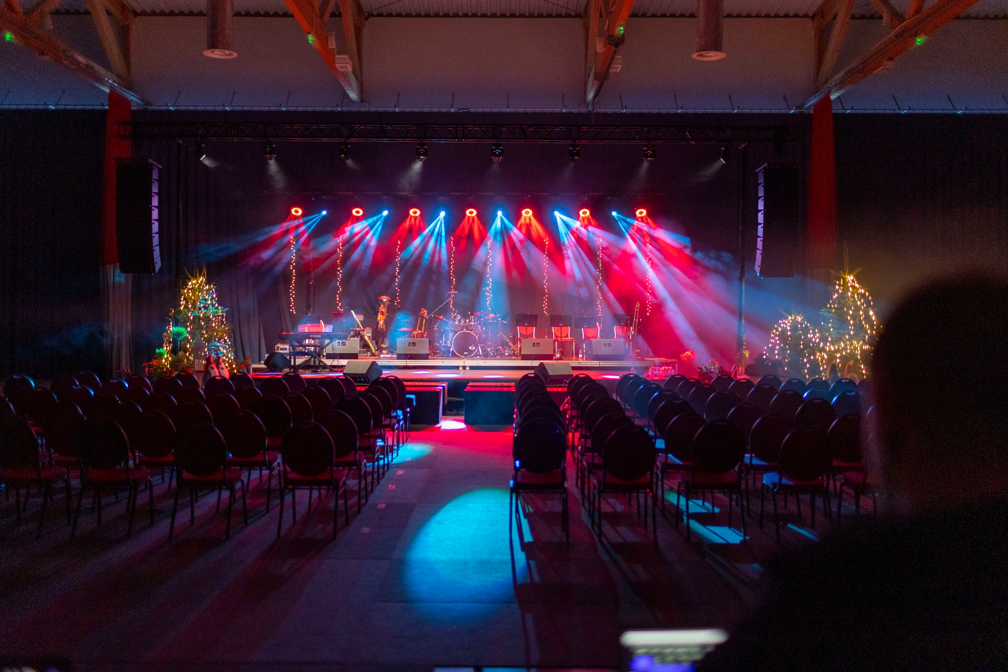 Scena przygotowana do występu z instrumentami, perkusją, rzędami krzeseł dla publiczności i dekoracjami świątecznymi, oświetlona czerwonymi i niebieskimi reflektorami.