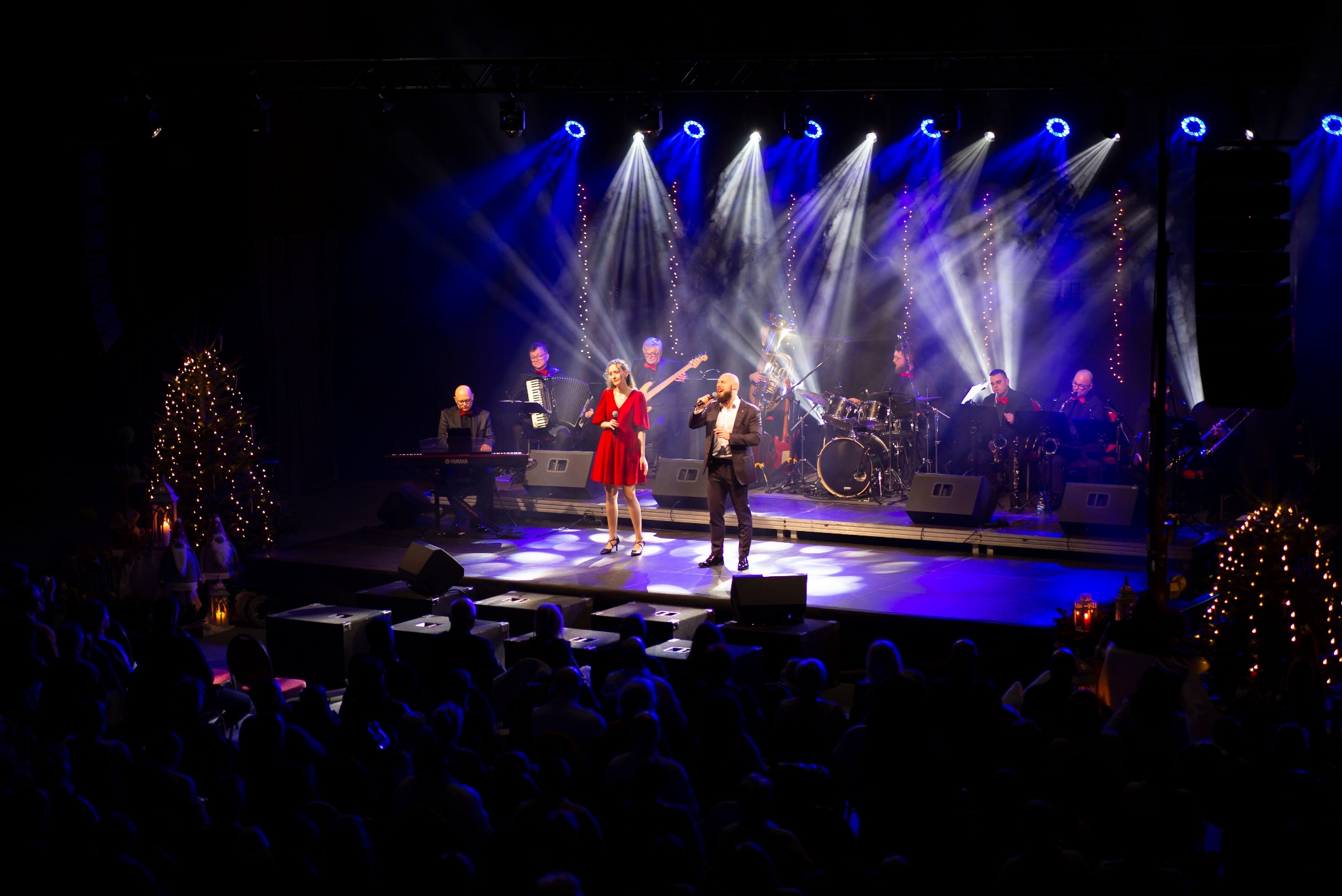 Duet wokalny na scenie z pełnym zespołem muzycznym, oświetlony reflektorami, widok z publicznością w tle.