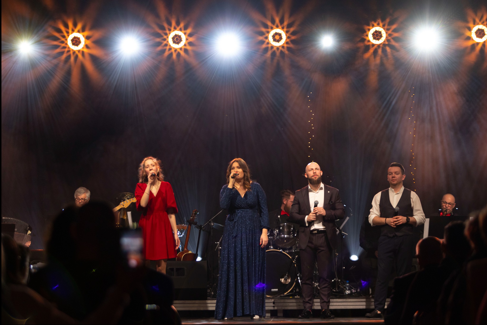 Wokalistki i wokaliści na scenie w blasku reflektorów, w tle zespół muzyczny i publiczność.