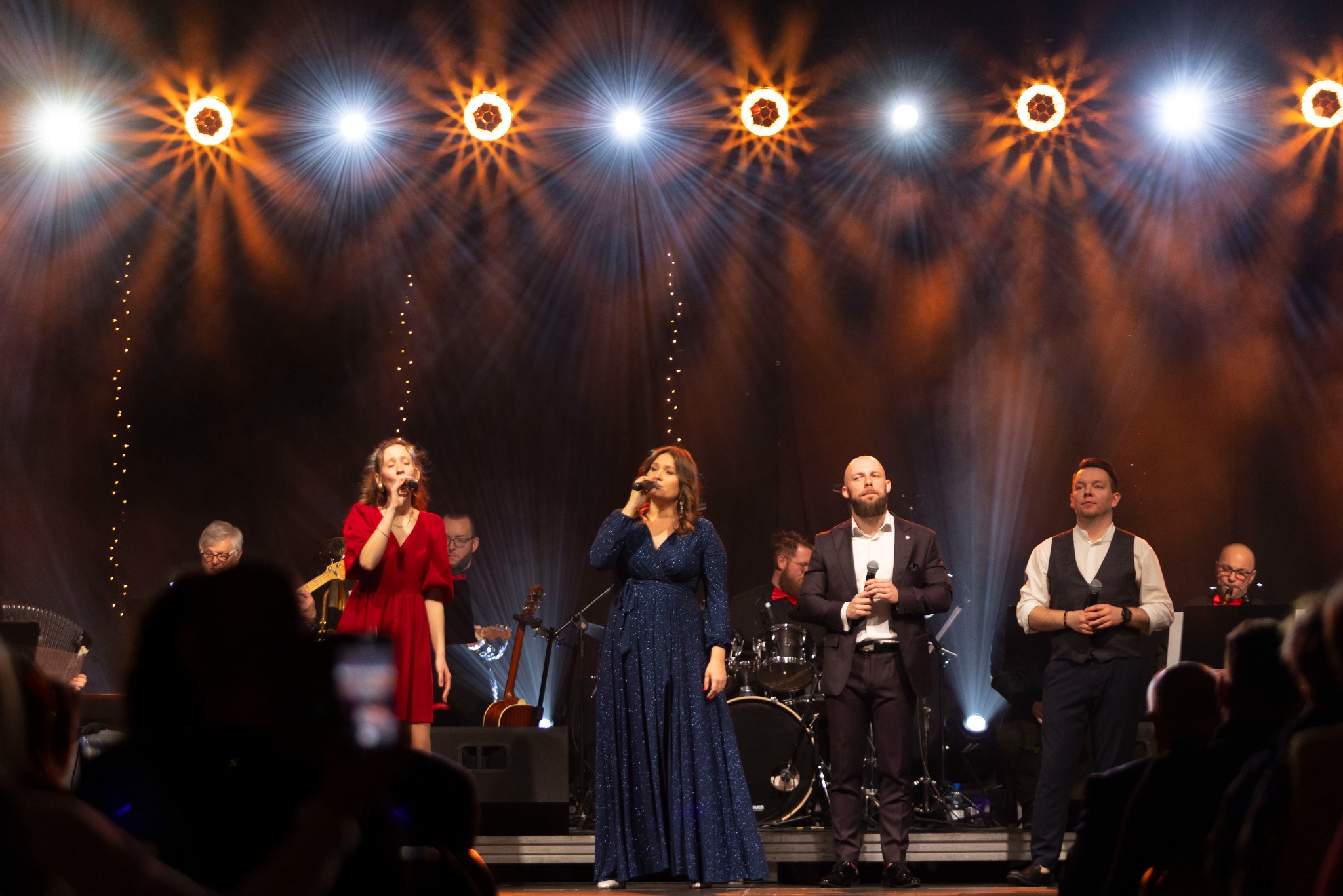 Zespół muzyczny na scenie podczas występu, oświetlony reflektorami, wokalistki w sukniach i muzycy w strojach galowych.