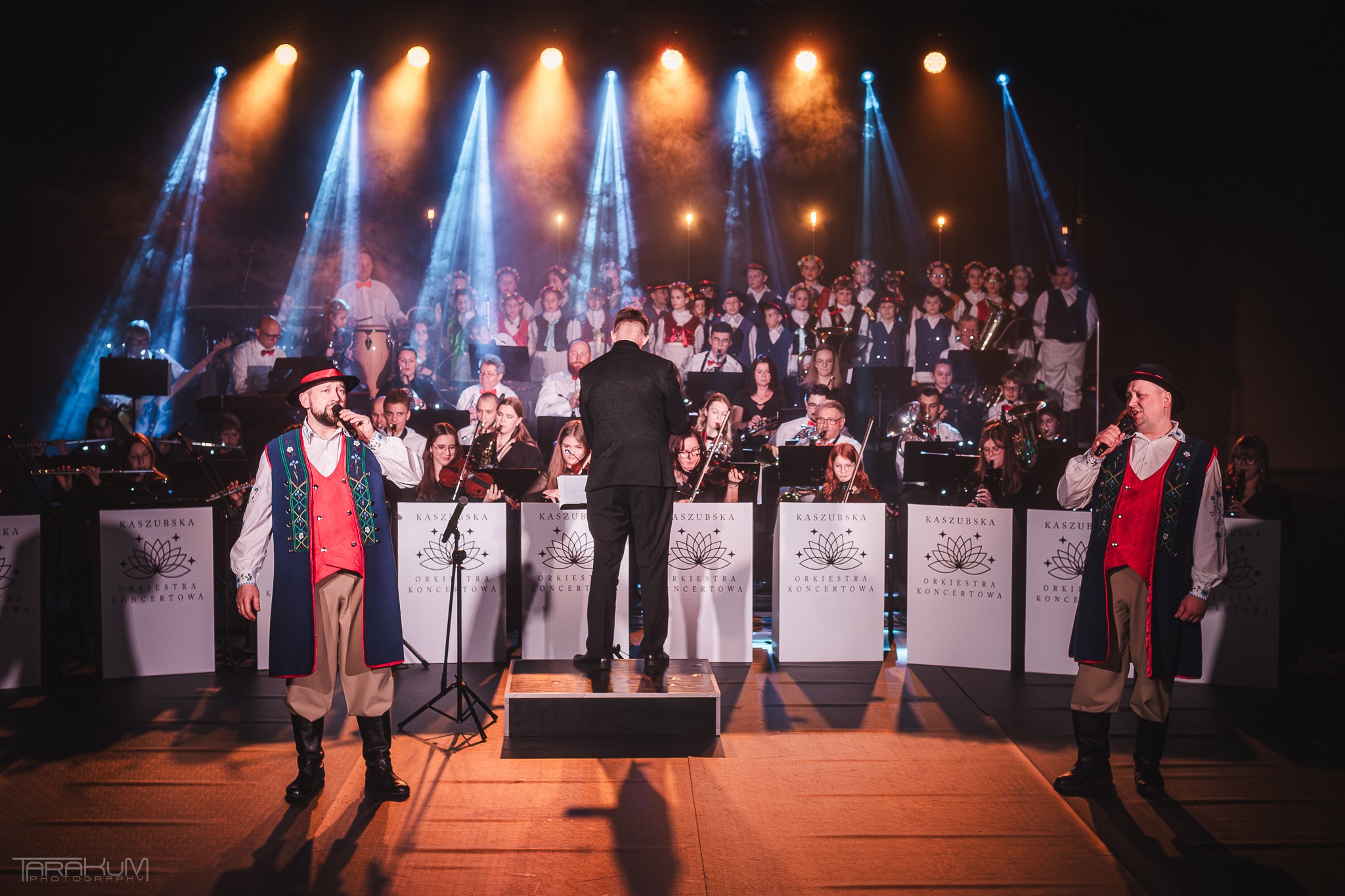 Orkiestra koncertowa w strojach ludowych, podświetlona reflektorami, dyrygent tyłem na podeście, scena z logotypami 'Kaszubska Orkiestra Koncertowa'.