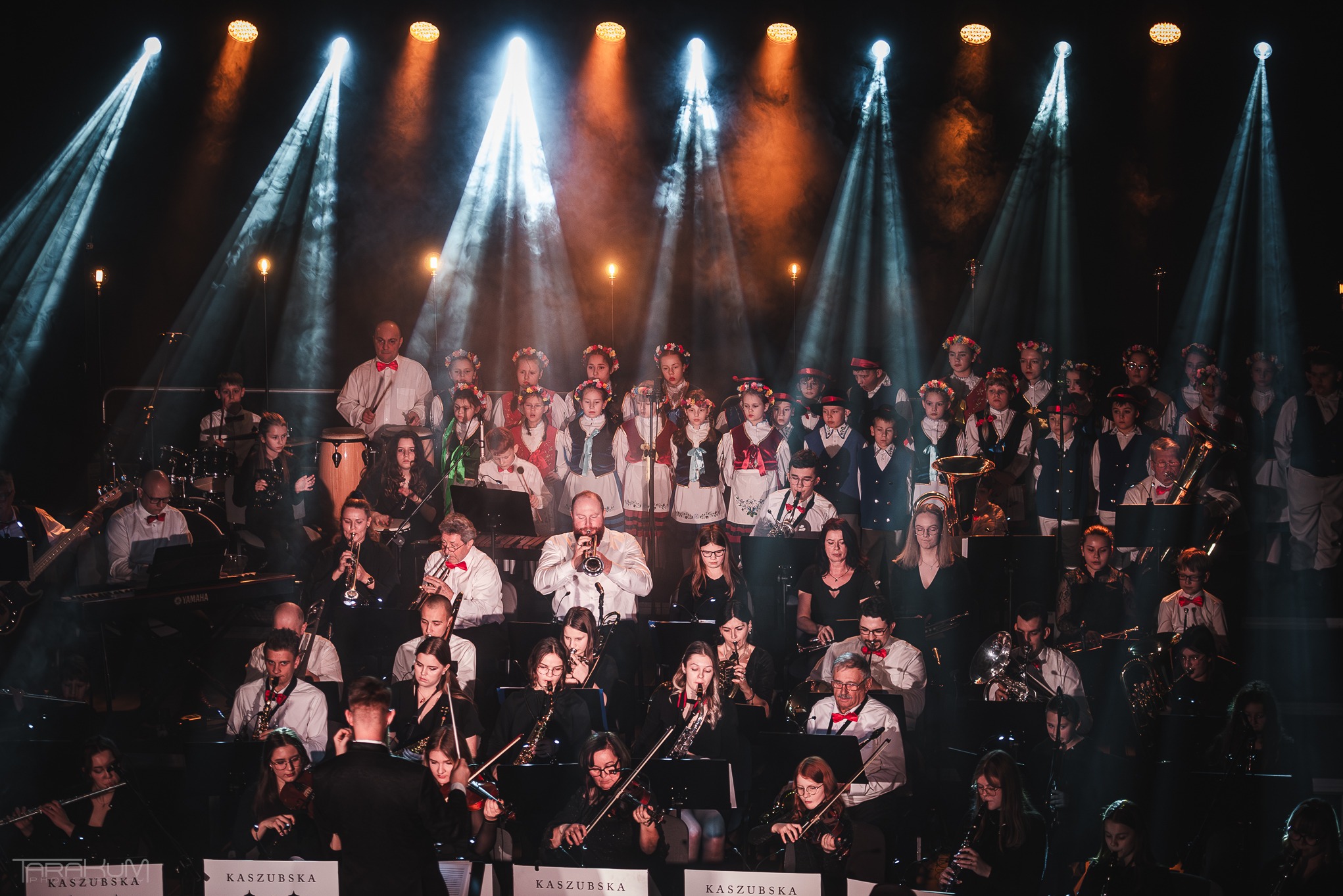 Duży zespół muzyczny z dyrygentem na scenie, oświetlony reflektorami, część osób w strojach ludowych.