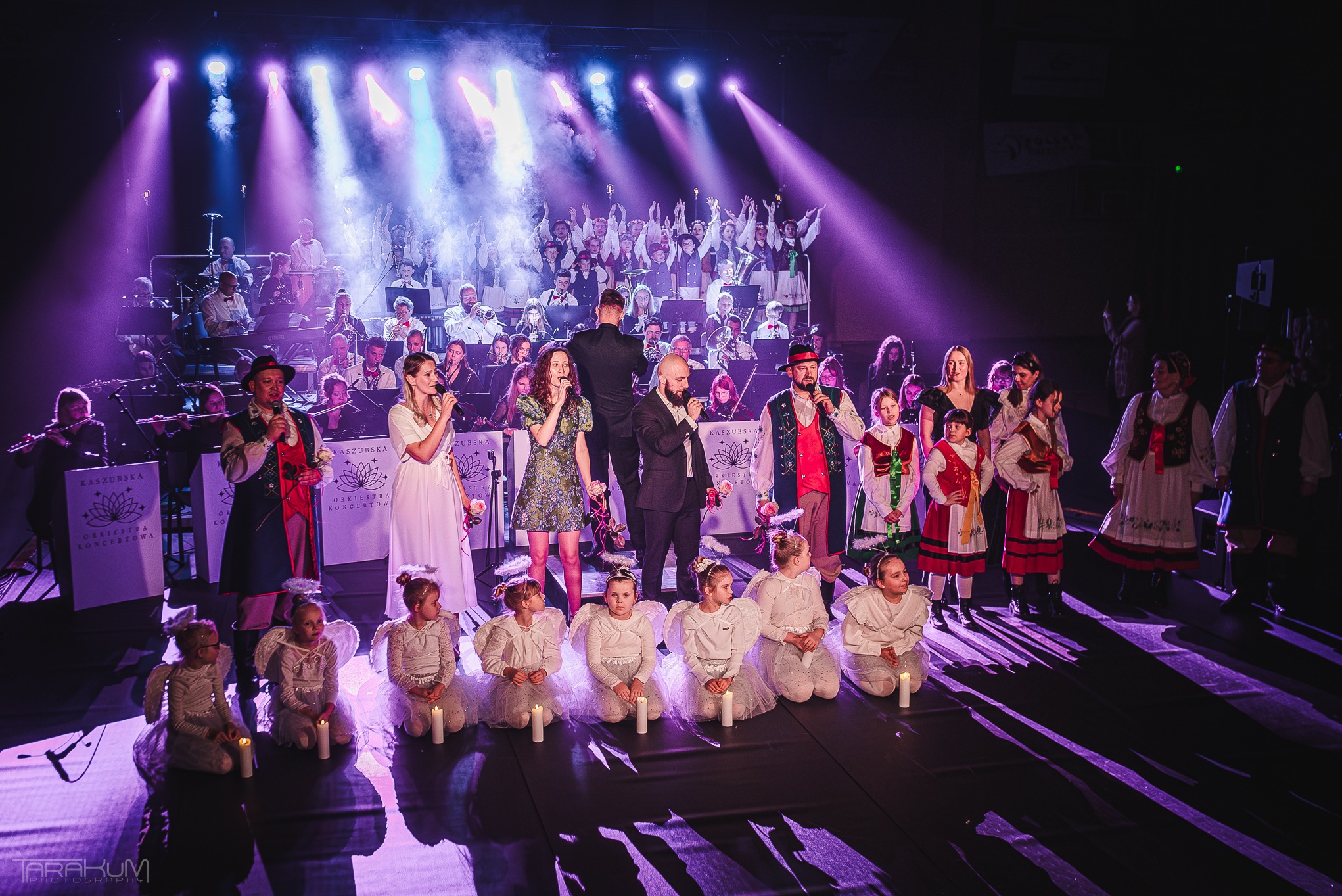 Scena z występu artystycznego: orkiestra, wokaliści w strojach ludowych i dzieci przebrane za anioły z zapalonymi świecami, oświetlona fioletowymi reflektorami.