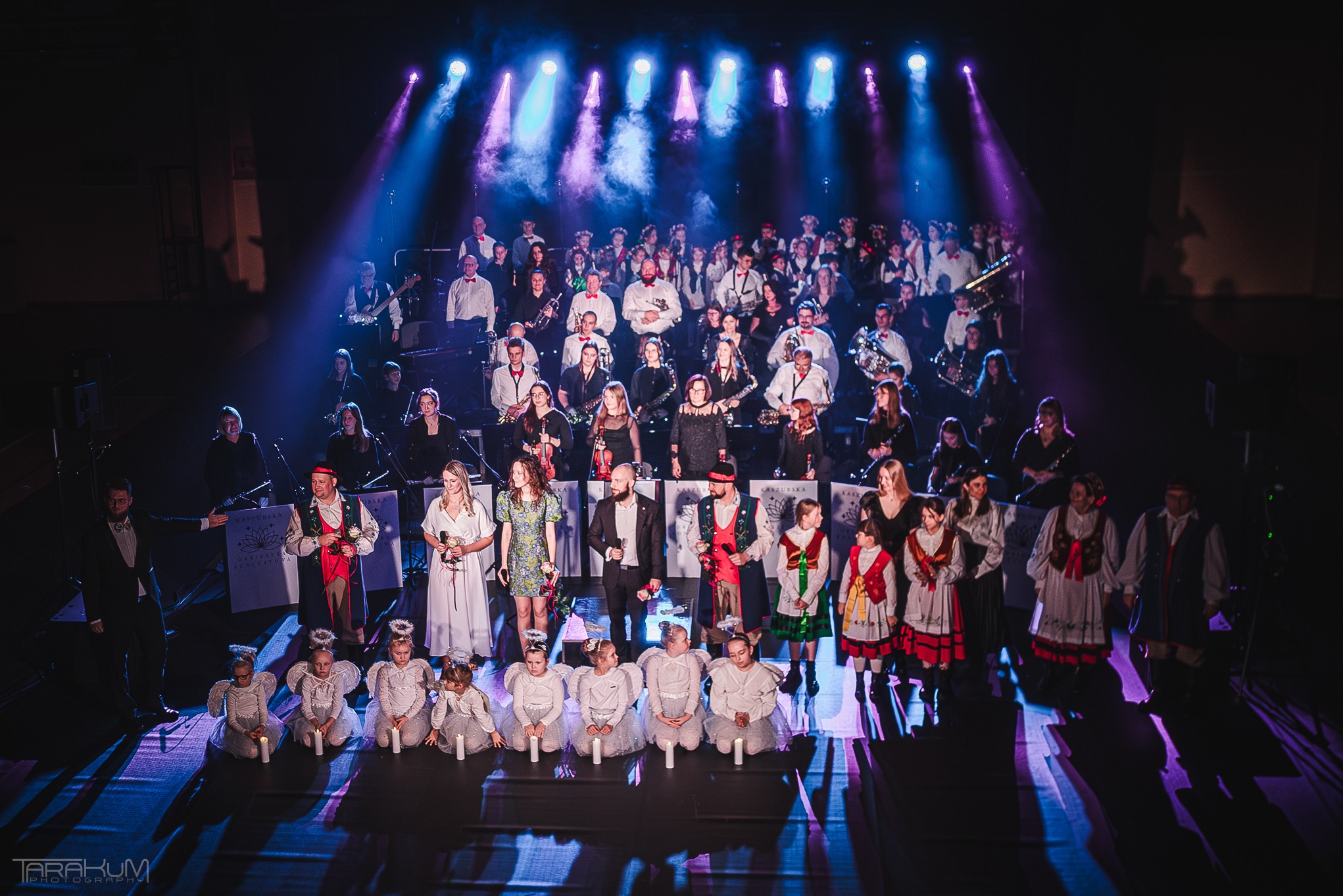 Duża grupa muzyków i dzieci w strojach aniołów na scenie oświetlonej reflektorami podczas występu.