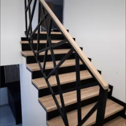 Nowoczesne schody z drewnianymi stopniami i czarną, geometryczną balustradą, jasne drewno poręczy, białe ściany w tle.