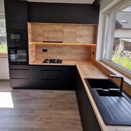 Mystictree - Nowoczesna kuchnia z czarnymi szafkami i drewnianymi blatami, oświetlenie LED pod szafkami górnymi, czarny zlew i bateria przy dużym oknie z widokiem na ogród.