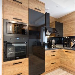 Mystictree - Nowoczesna kuchnia z drewnianymi frontami szafek i czarnymi, błyszczącymi elementami, zabudowany piekarnik i mikrofalówka marki Samsung, czarny okap i blat z ciemnego kamienia.