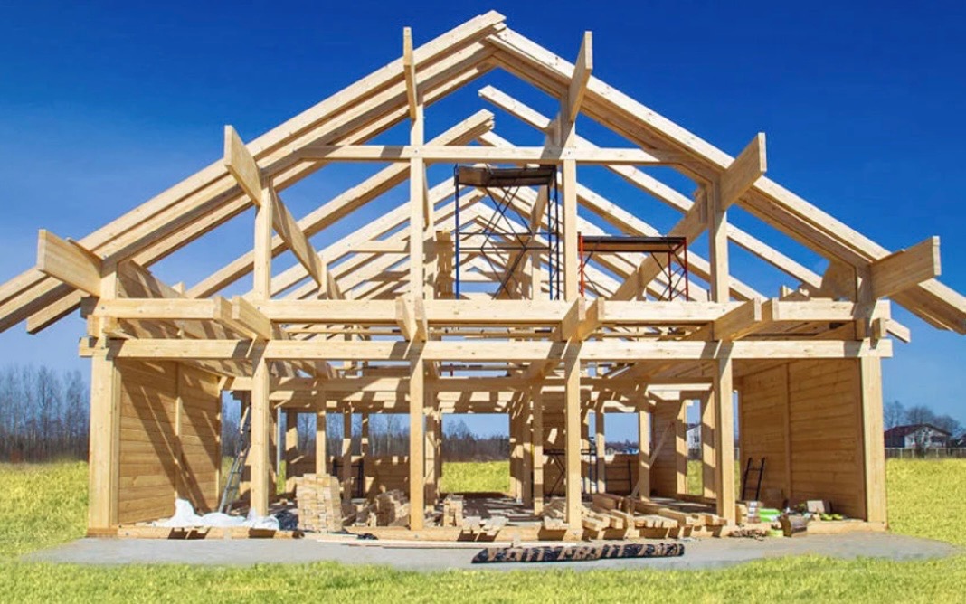Konstrukcja drewniana domu w trakcie budowy, widoczne rusztowania, elementy ścian z bali, jasne drewno na tle błękitnego nieba.