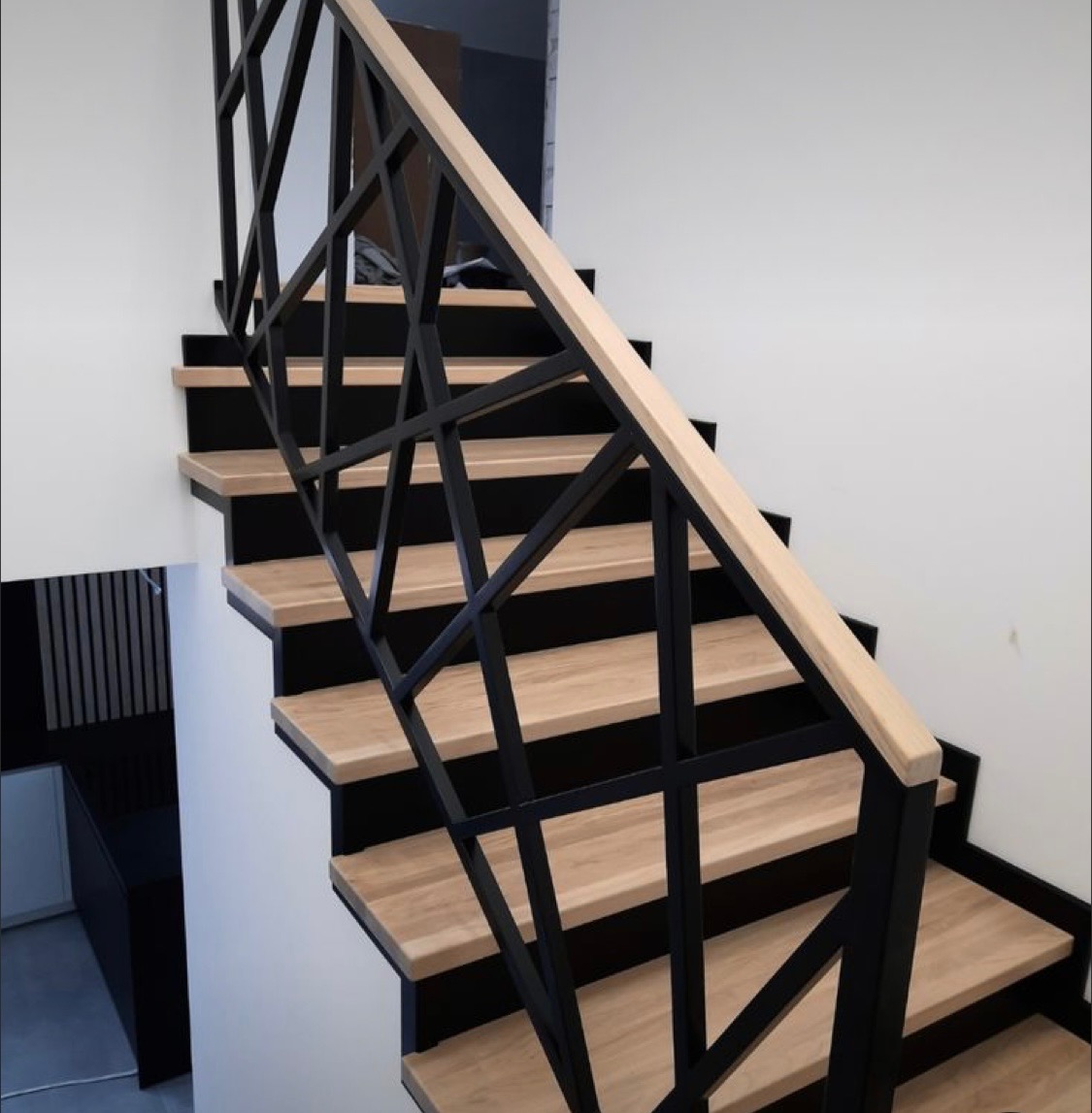 Nowoczesne schody z drewnianymi stopniami i czarną, geometryczną balustradą, jasne drewno poręczy, białe ściany w tle.