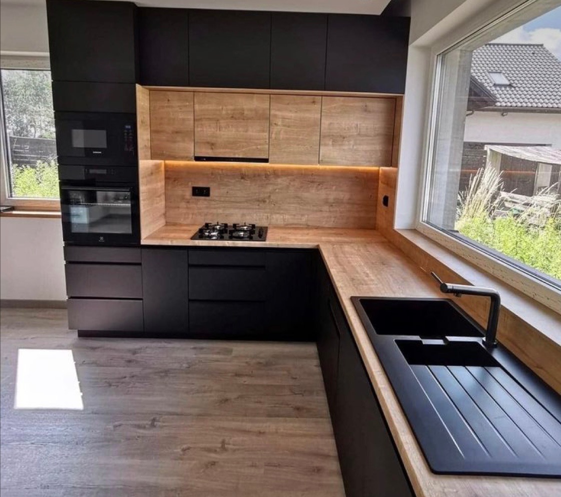 Nowoczesna kuchnia z czarnymi szafkami i drewnianymi blatami, oświetlenie LED pod szafkami górnymi, czarny zlew i bateria przy dużym oknie z widokiem na ogród.