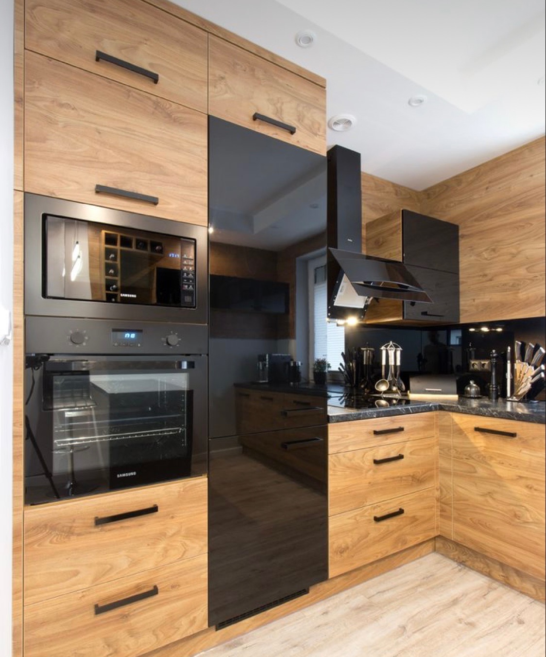 Nowoczesna kuchnia z drewnianymi frontami szafek i czarnymi, błyszczącymi elementami, zabudowany piekarnik i mikrofalówka marki Samsung, czarny okap i blat z ciemnego kamienia.