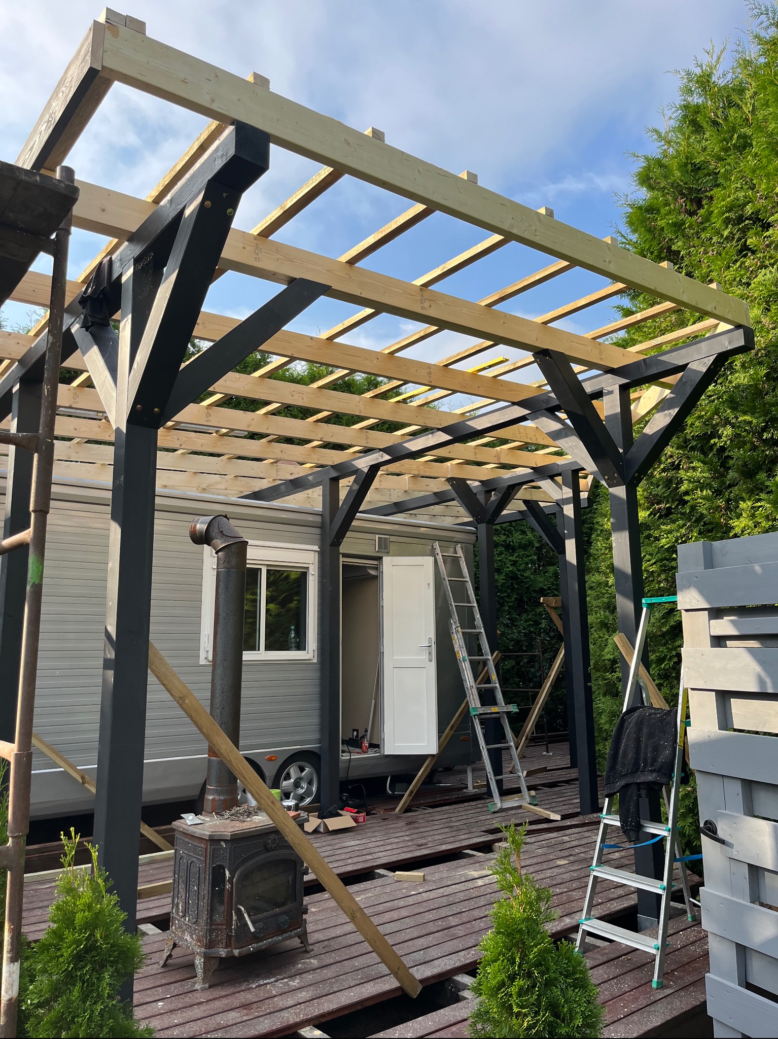 Konstrukcja z belek: czarne słupy podtrzymują surowy szkielet dachu z jasnego drewna, obok przyczepa kempingowa i piec kominkowy, na podłodze z desek porozrzucane narzędzia i resztki materiałów.