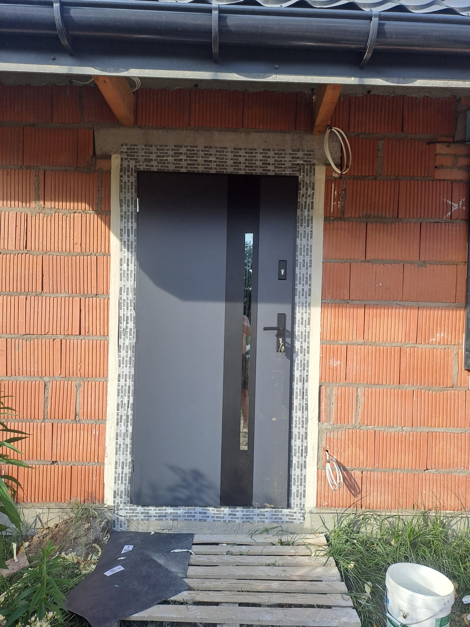 Szare drzwi wejściowe z przeszkleniem, zamontowane w surowym murze z cegły, zabezpieczone taśmą okienną, z tymczasowym podejściem z palet i wiadrem farby.