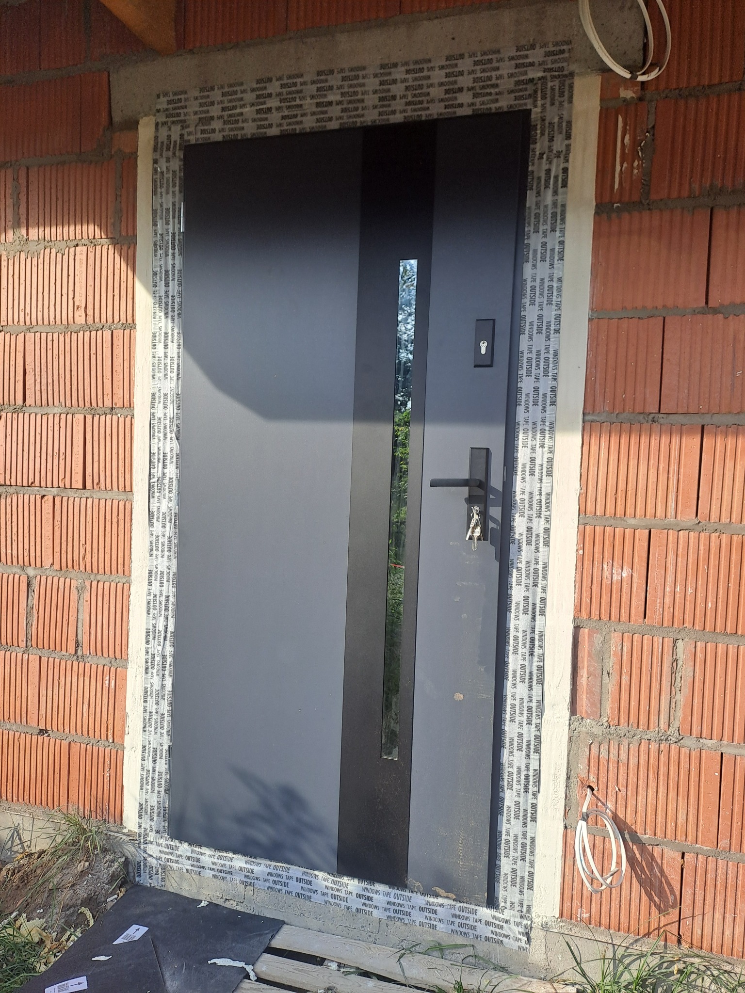 Montaż nowoczesnych, grafitowych drzwi wejściowych z wąskim przeszkleniem, zabezpieczonych taśmą uszczelniającą na surowej ścianie z czerwonej cegły.