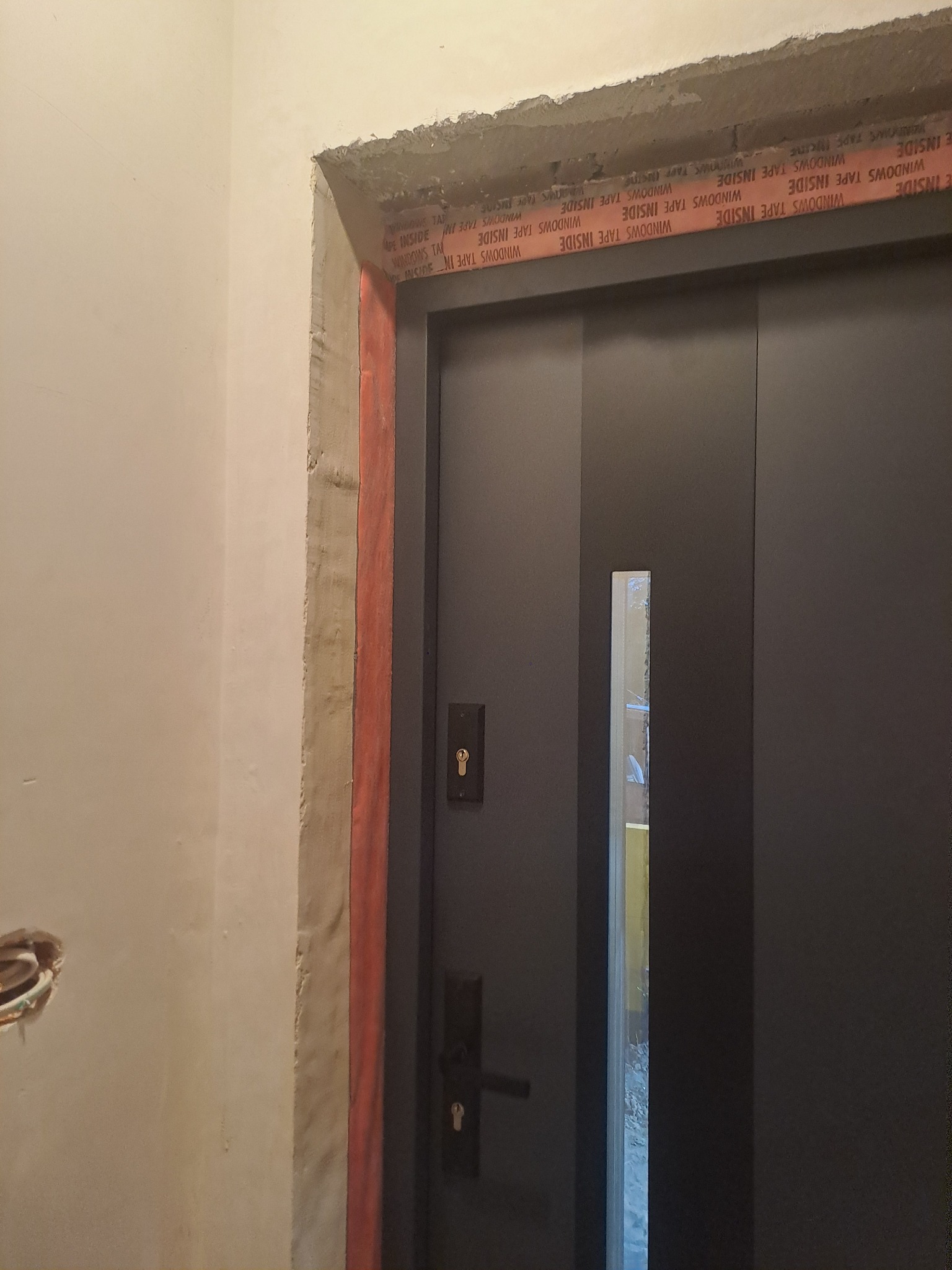 Ciemne drzwi wejściowe z wąskim przeszkleniem, widoczne ślady prac montażowych: pianka montażowa i taśma uszczelniająca 'WINDOWS TAPE INSIDE' wokół ościeżnicy na surowej ścianie.