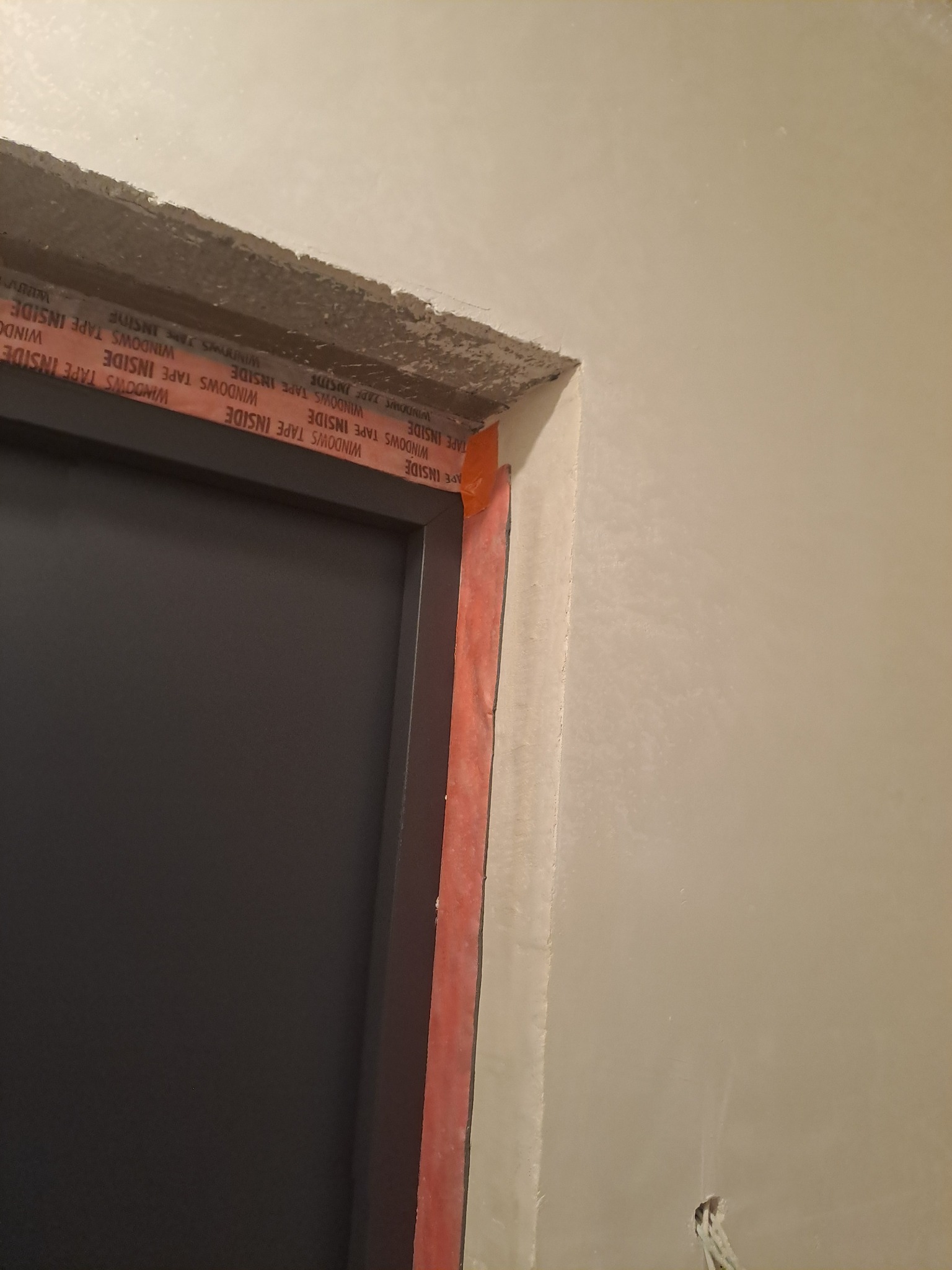 Fragment ościeżnicy z zamontowaną czarną ramą drzwiową, zabezpieczoną czerwoną taśmą uszczelniającą 'WINDOWS TAPE INSIDE', widoczne surowe wykończenie ściany i betonu nad drzwiami.