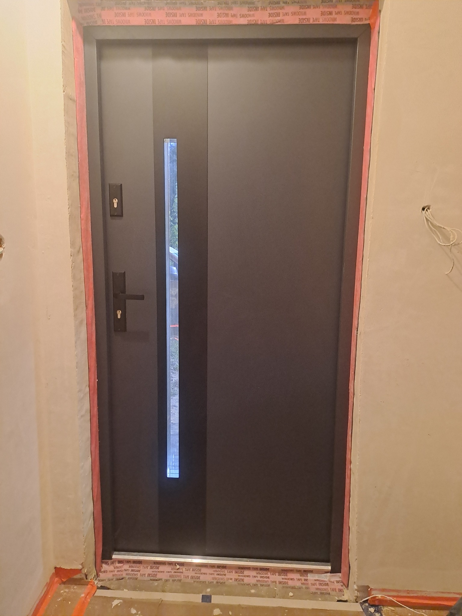 Szare drzwi wejściowe z długim, wąskim przeszkleniem, zabezpieczone taśmą uszczelniającą na styku z murem w trakcie montażu, widoczne przewody elektryczne