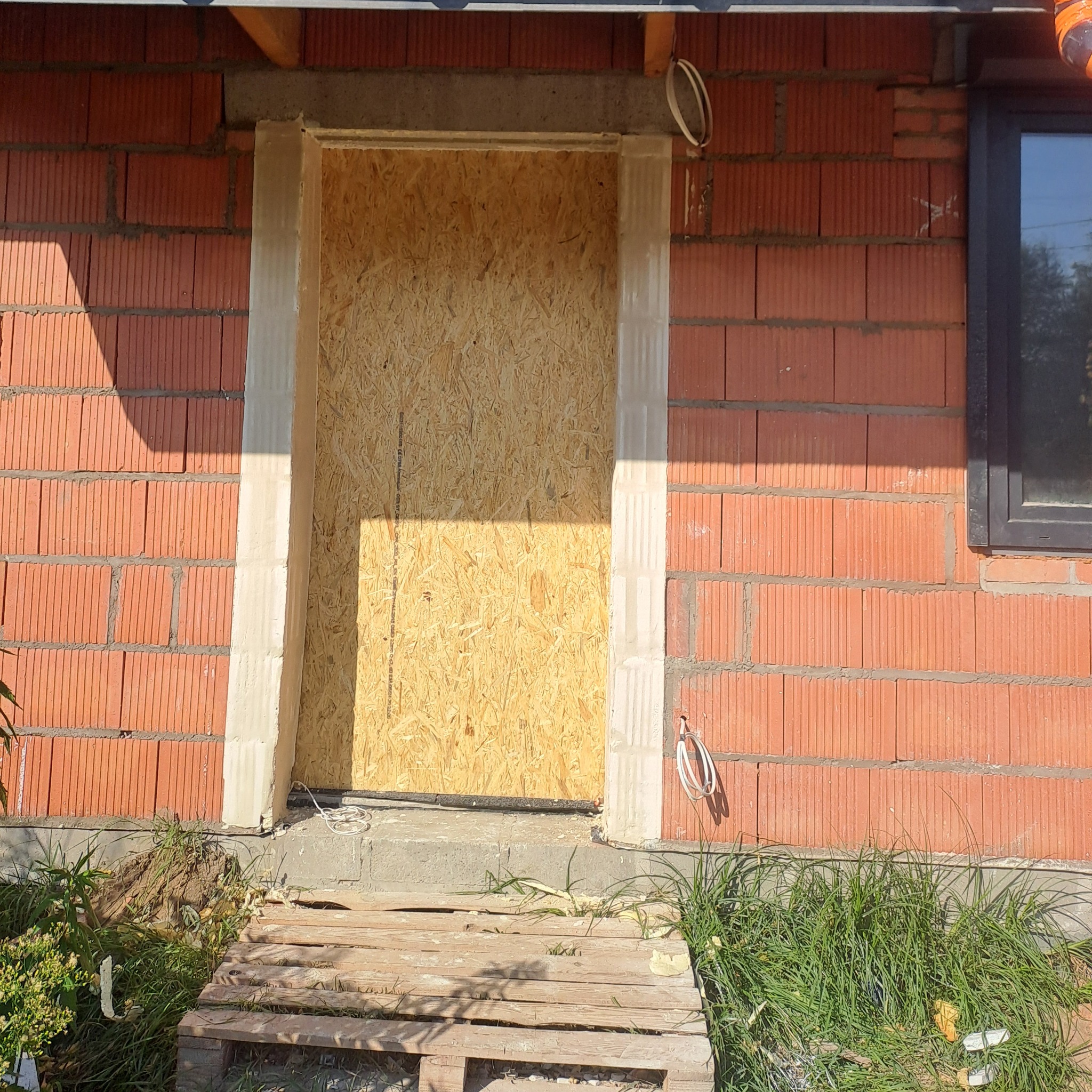 Surowy otwór drzwiowy w bloku z czerwonej cegły, zabezpieczony płytą OSB, z widocznymi przewodami elektrycznymi i paletą jako prowizorycznym wejściem.