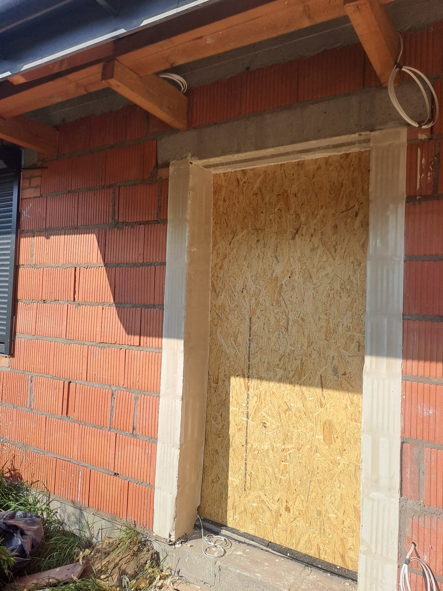 Surowy otwór drzwiowy w ceglanym murze, zabezpieczony płytą OSB, widoczne świeże obramowanie z betonu komórkowego i wystające przewody elektryczne.