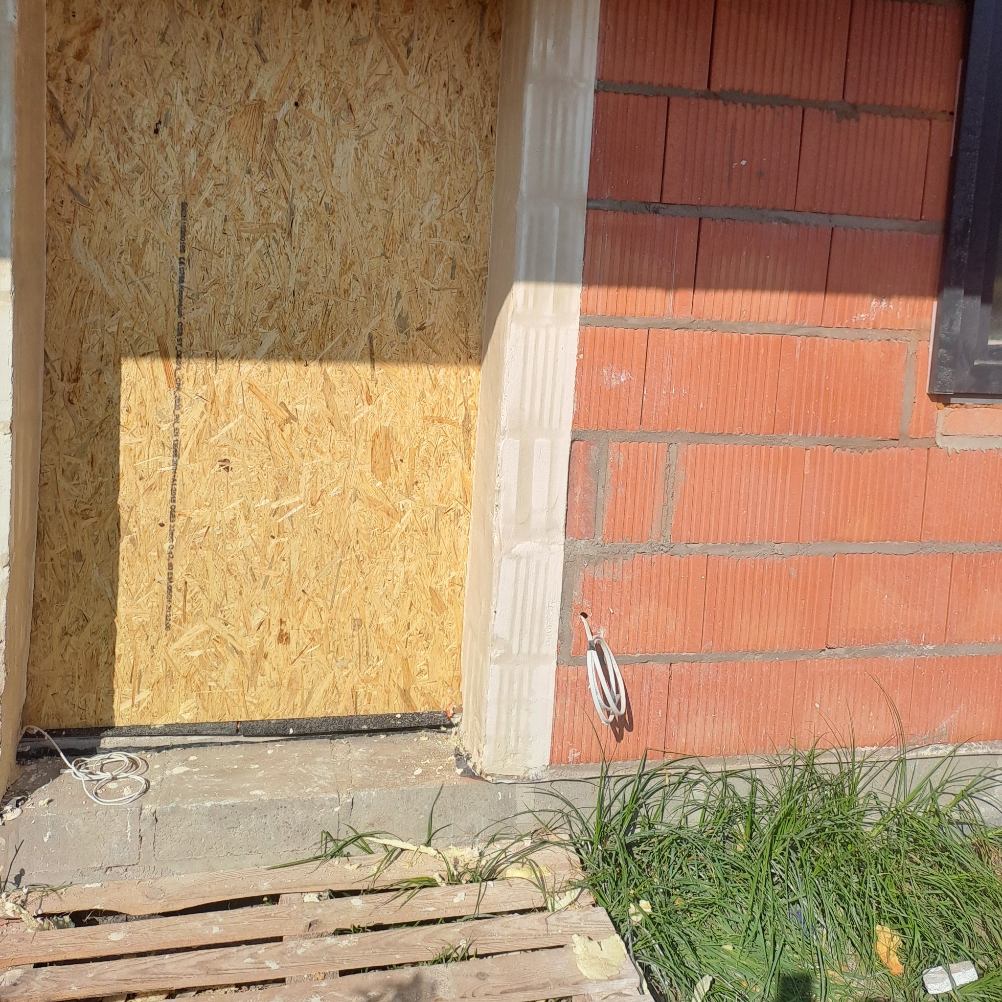 Zabezpieczony otwór drzwiowy sklejką OSB w trakcie prac budowlanych, widoczny fragment ściany z czerwonej cegły i wystające przewody.