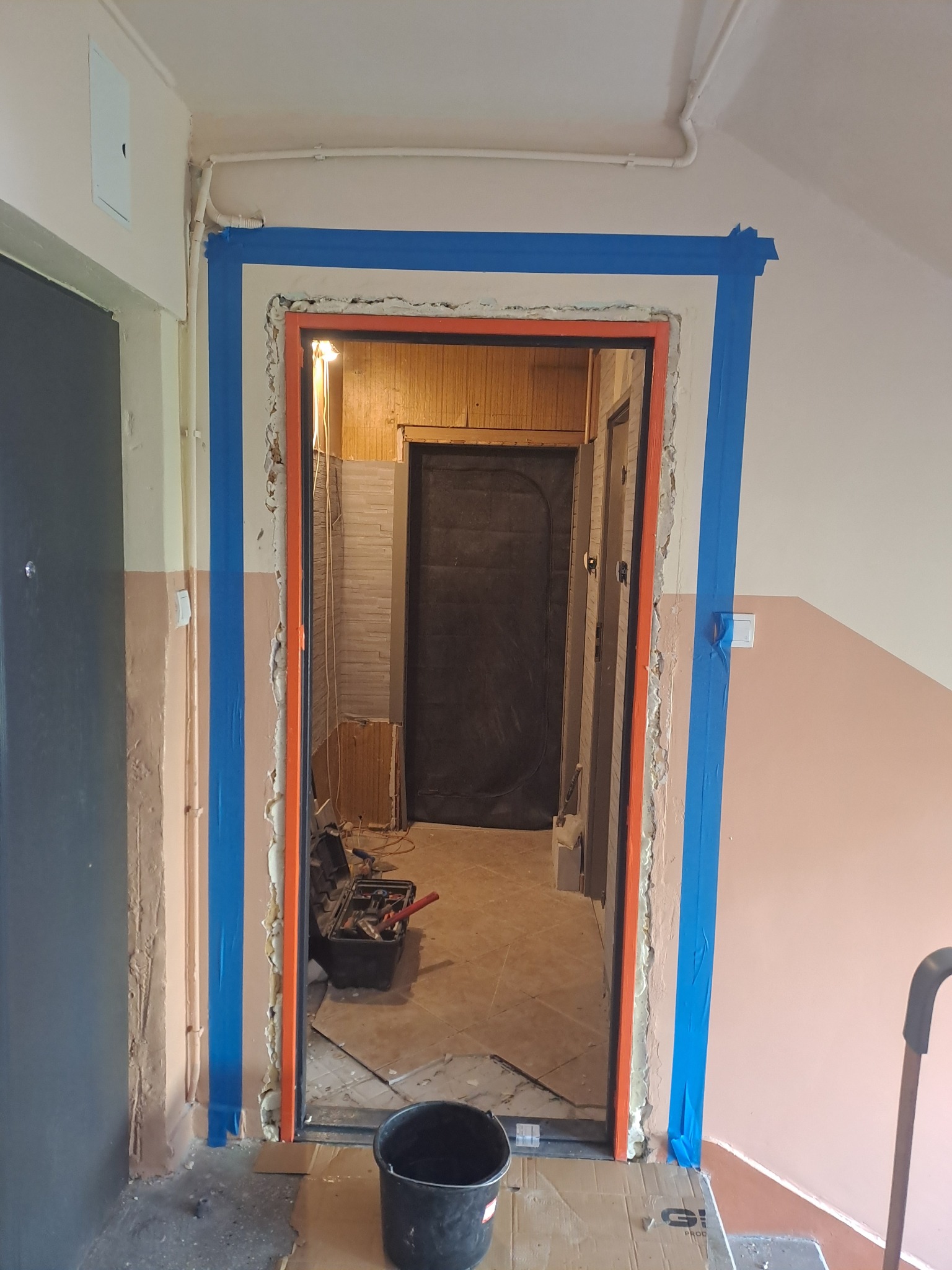 Przygotowane do osadzenia ościeżnicy wejście, zabezpieczone niebieską taśmą malarską, z widoczną pianką montażową i narzędziami w tle.