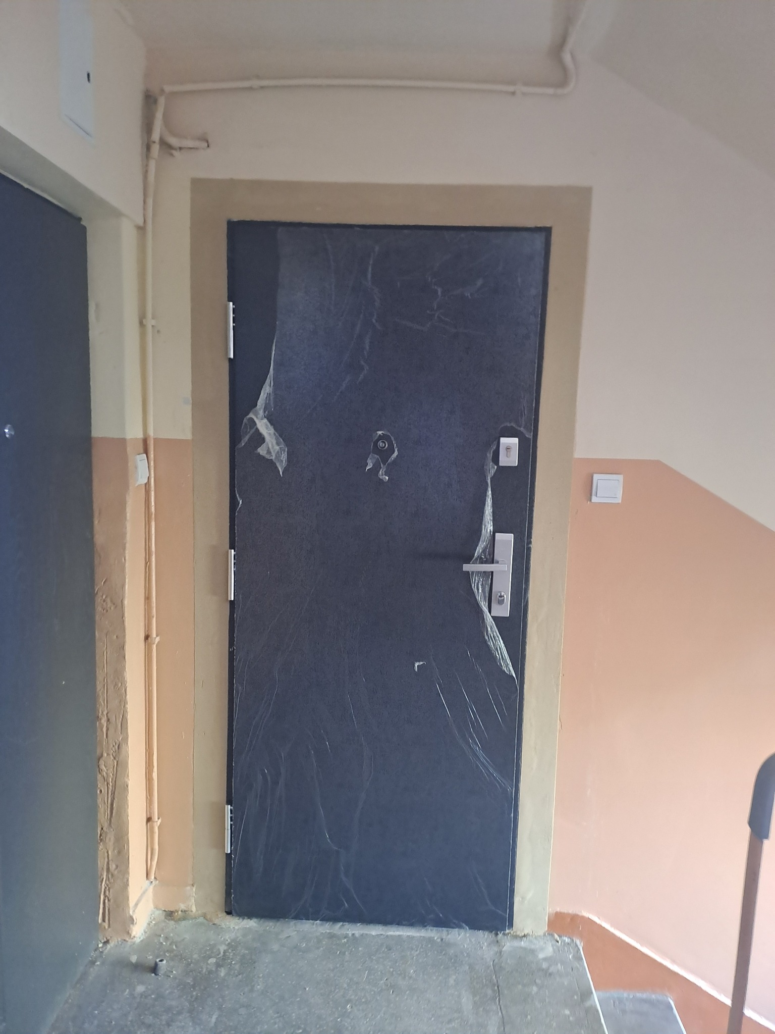 Nowo zamontowane, ciemne drzwi wejściowe z widoczną folią ochronną, osadzone w beżowej futrynie na tle klatki schodowej.