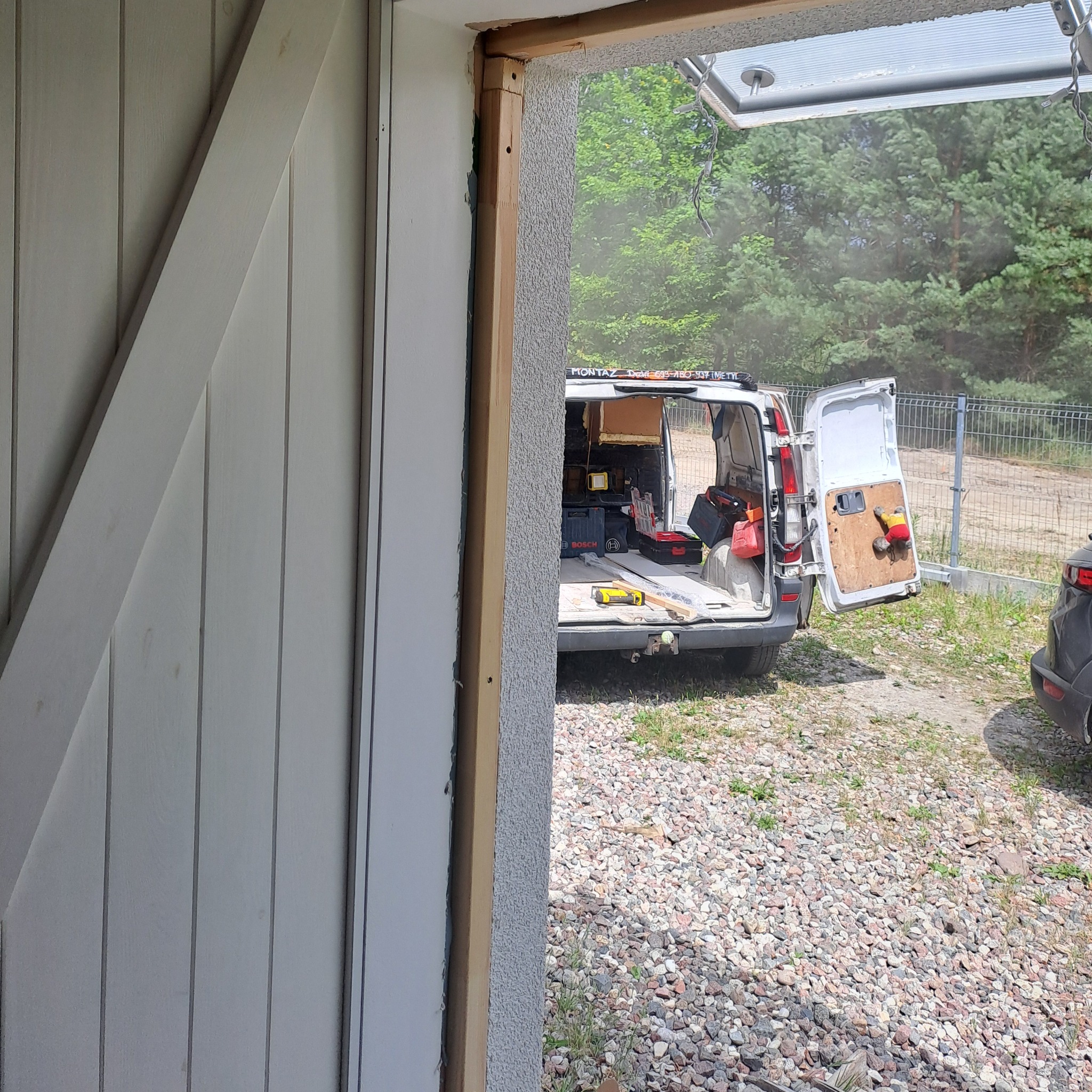 Rama drzwiowa z widocznym samochodem dostawczym z otwartymi drzwiami i narzędziami w tle, na zewnątrz żwirowe podłoże i zieleń.