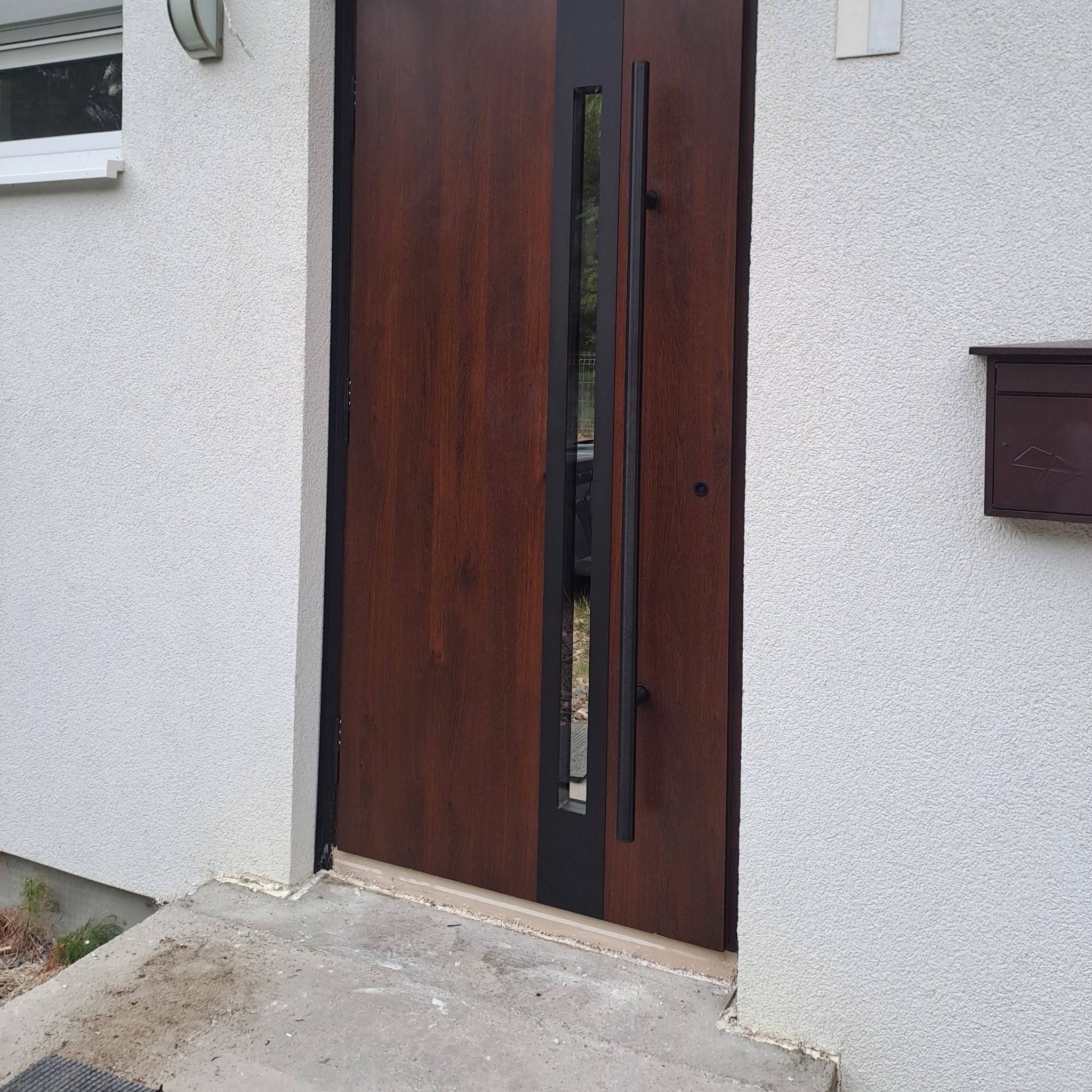 Nowoczesne, brązowe drzwi wejściowe z czarnymi elementami i długim uchwytem, zamontowane w domu w Łodzi, widoczne z zewnątrz, obok skrzynka pocztowa.