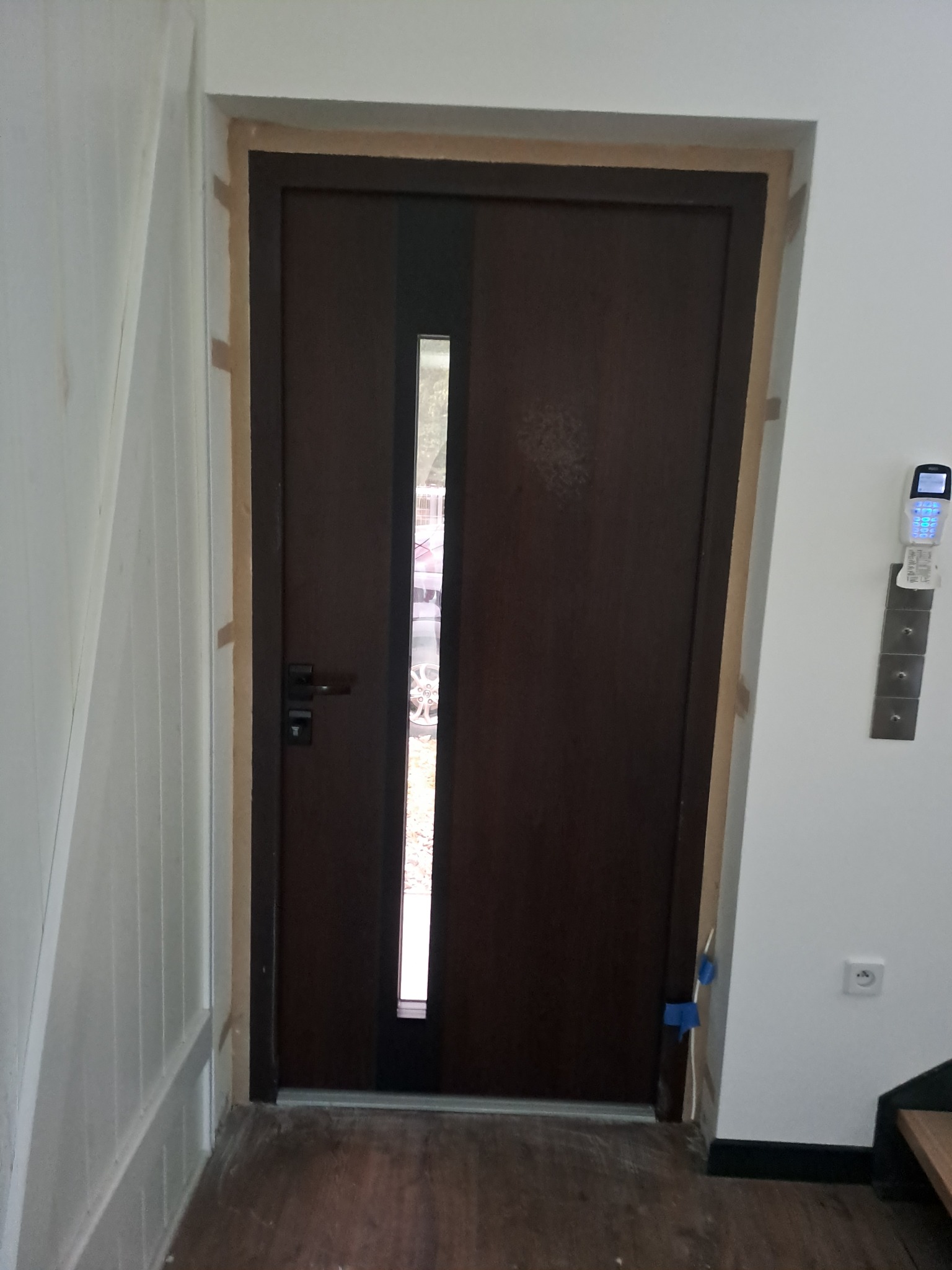 Ciemnobrązowe drzwi wejściowe z pionowym przeszkleniem, częściowo osadzone w ościeżnicy, widoczne niebieskie taśmy zabezpieczające i szczeliny montażowe przed wykończeniem.