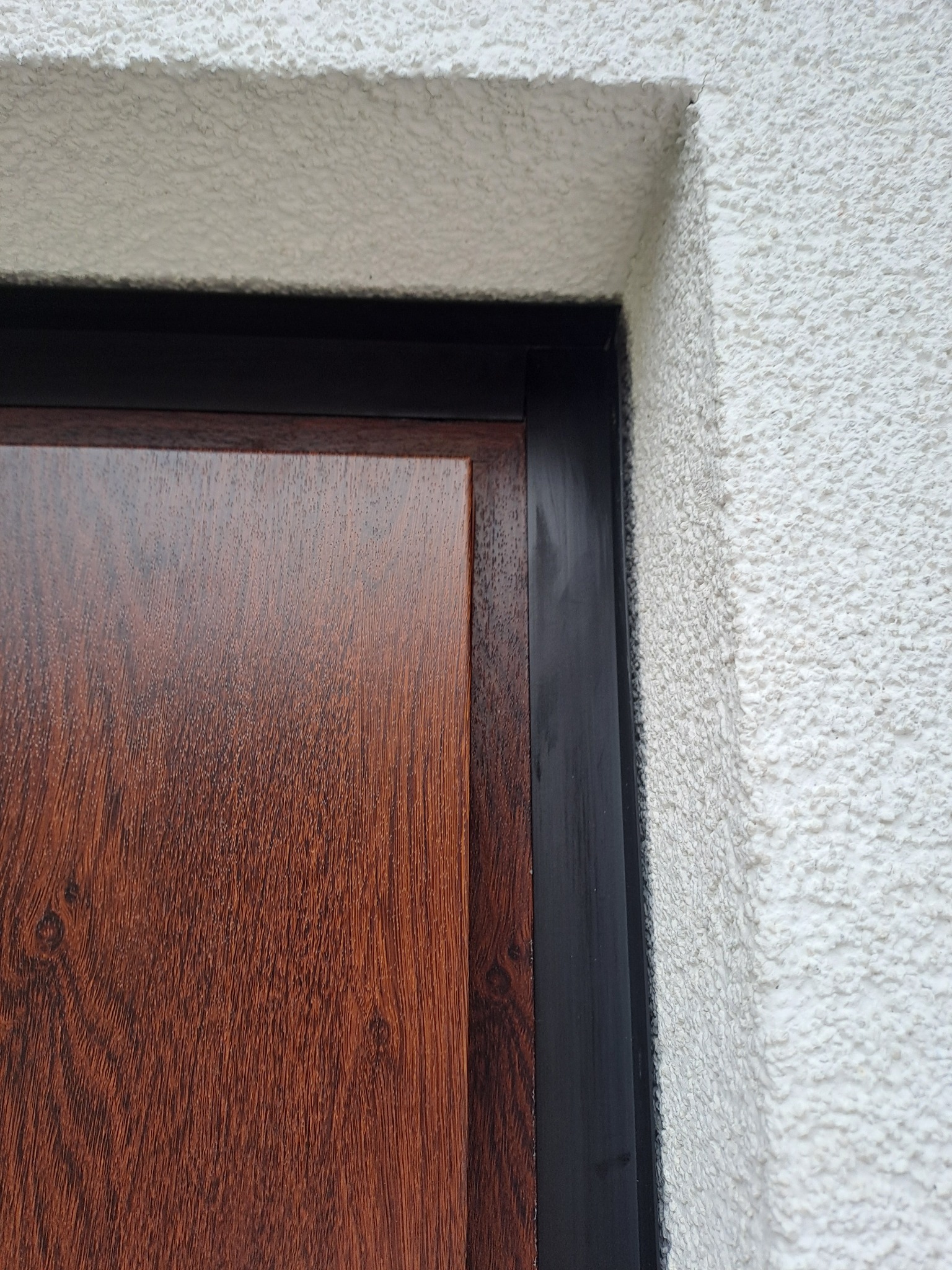 Fragment brązowych drzwi z czarną ościeżnicą, widoczna struktura białego tynku na ścianie zewnętrznej.