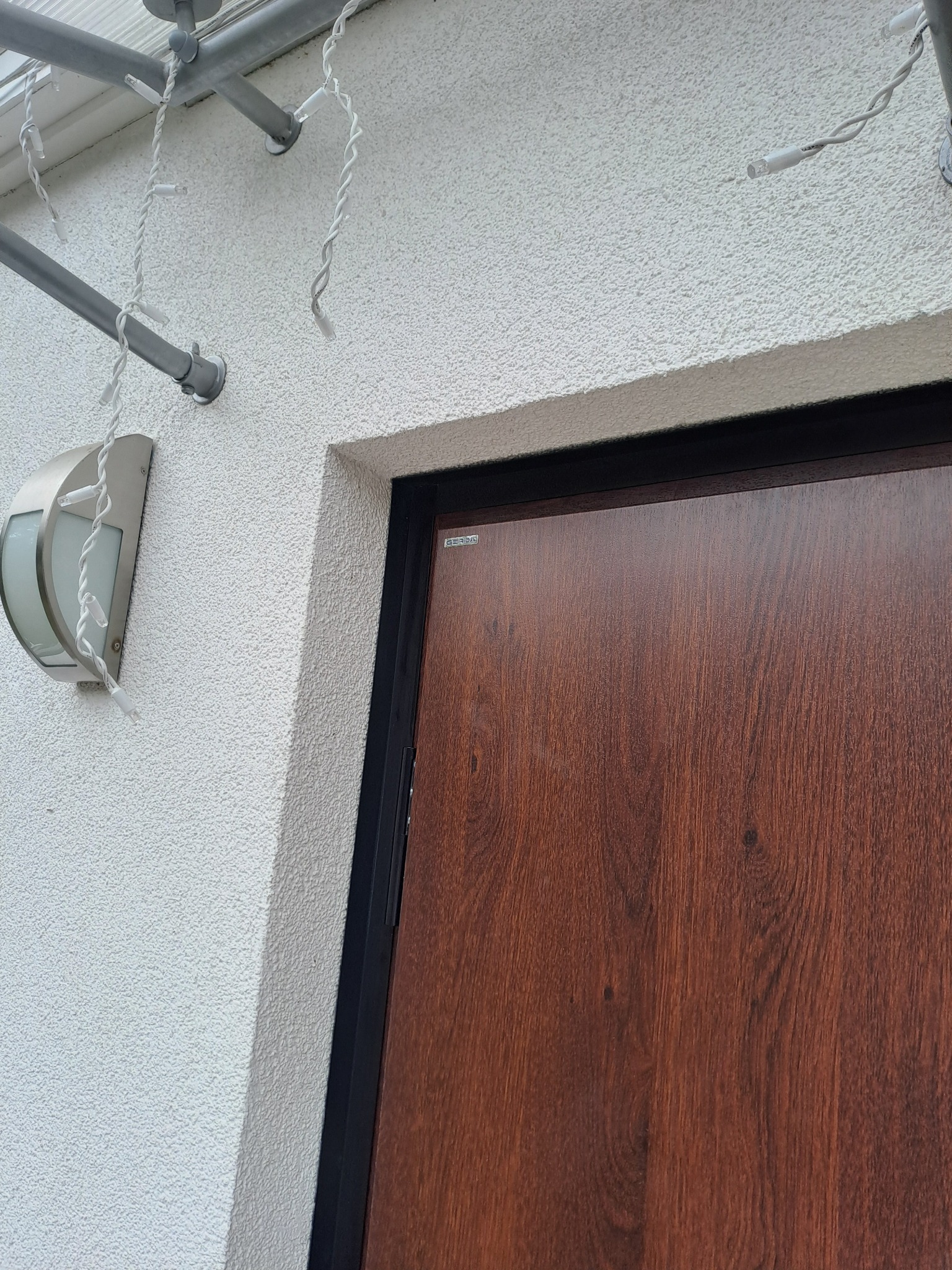 Ciemnobrązowe drzwi wejściowe z czarną ramą, oświetlone lampą elewacyjną, dekorowane białymi lampkami, na tle białej, chropowatej ściany.