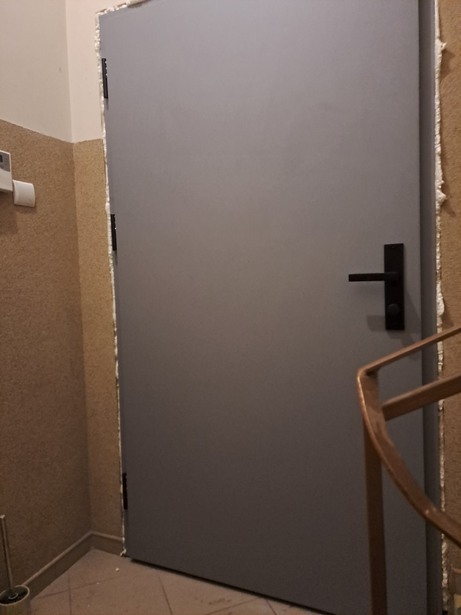 Szare drzwi wejściowe z czarną klamką, osadzone w ościeżnicy z widoczną pianką montażową i fragmentem drewnianej balustrady schodów w tle.