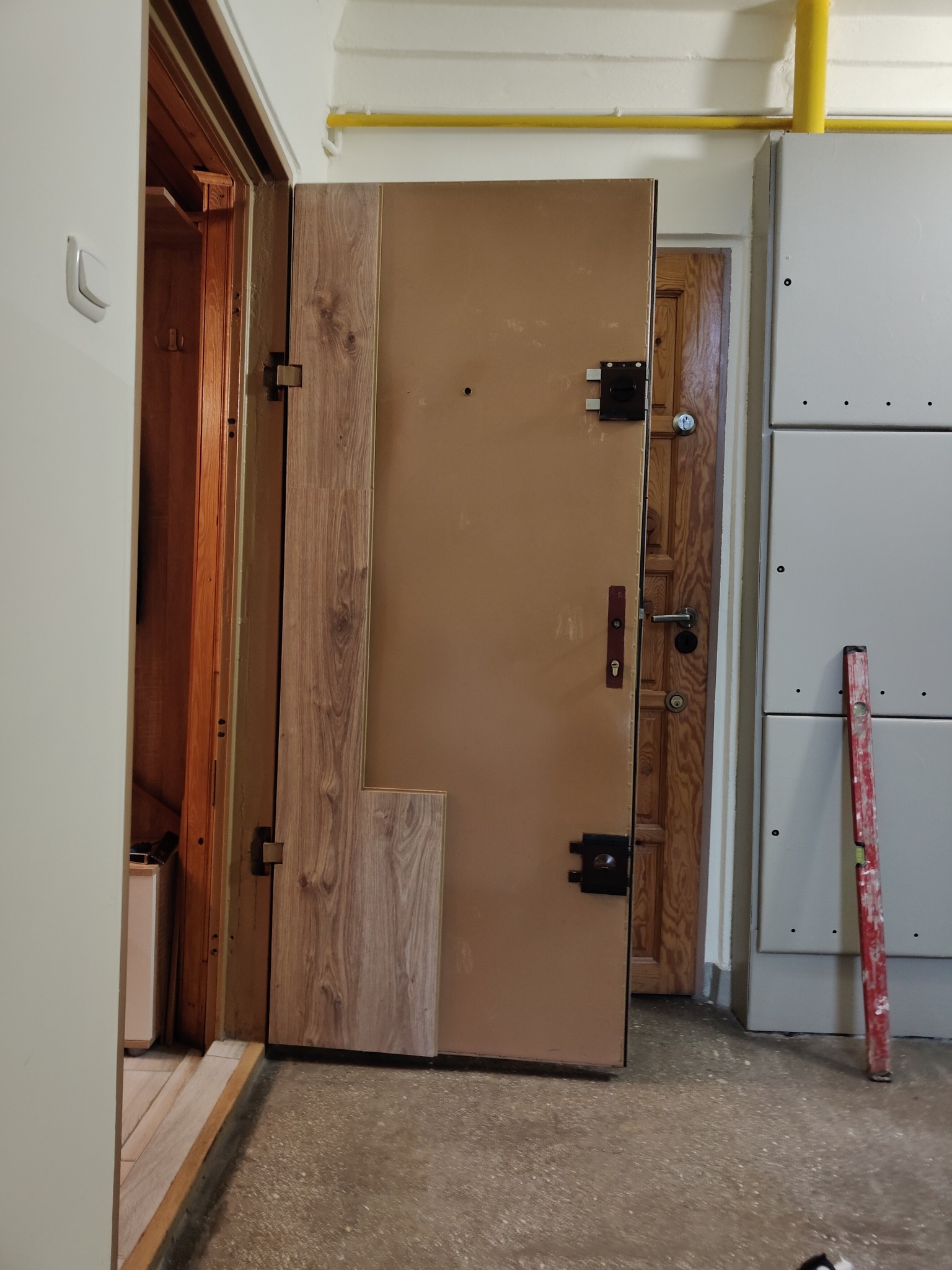 Brązowe drzwi wejściowe z fragmentem okleiny imitującej drewno, widoczny poziomica oparta o ścianę, w tle drewniane drzwi wewnętrzne.